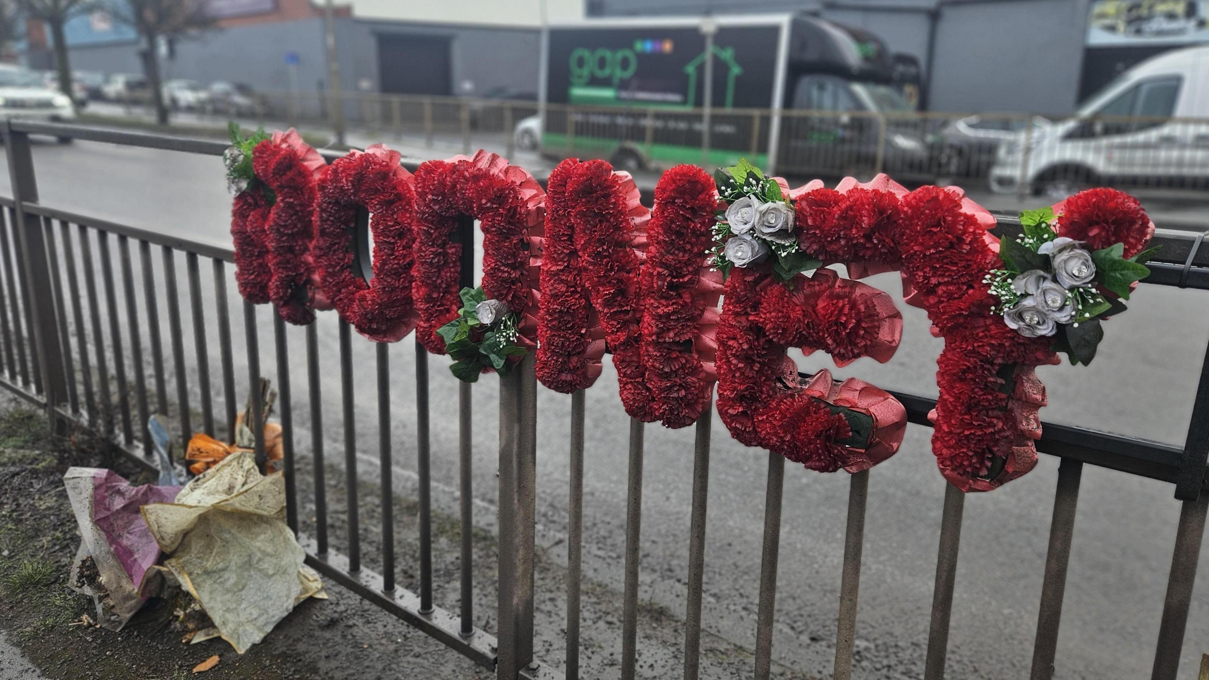 A metal crash barrier with a floral wreath attached to it. It's on a busy road with traffic and there are shop and office frontages in the background. The wreath is made of red flowers spelling out "Rodney". Two bouquets of flowers are also visible on the ground. 