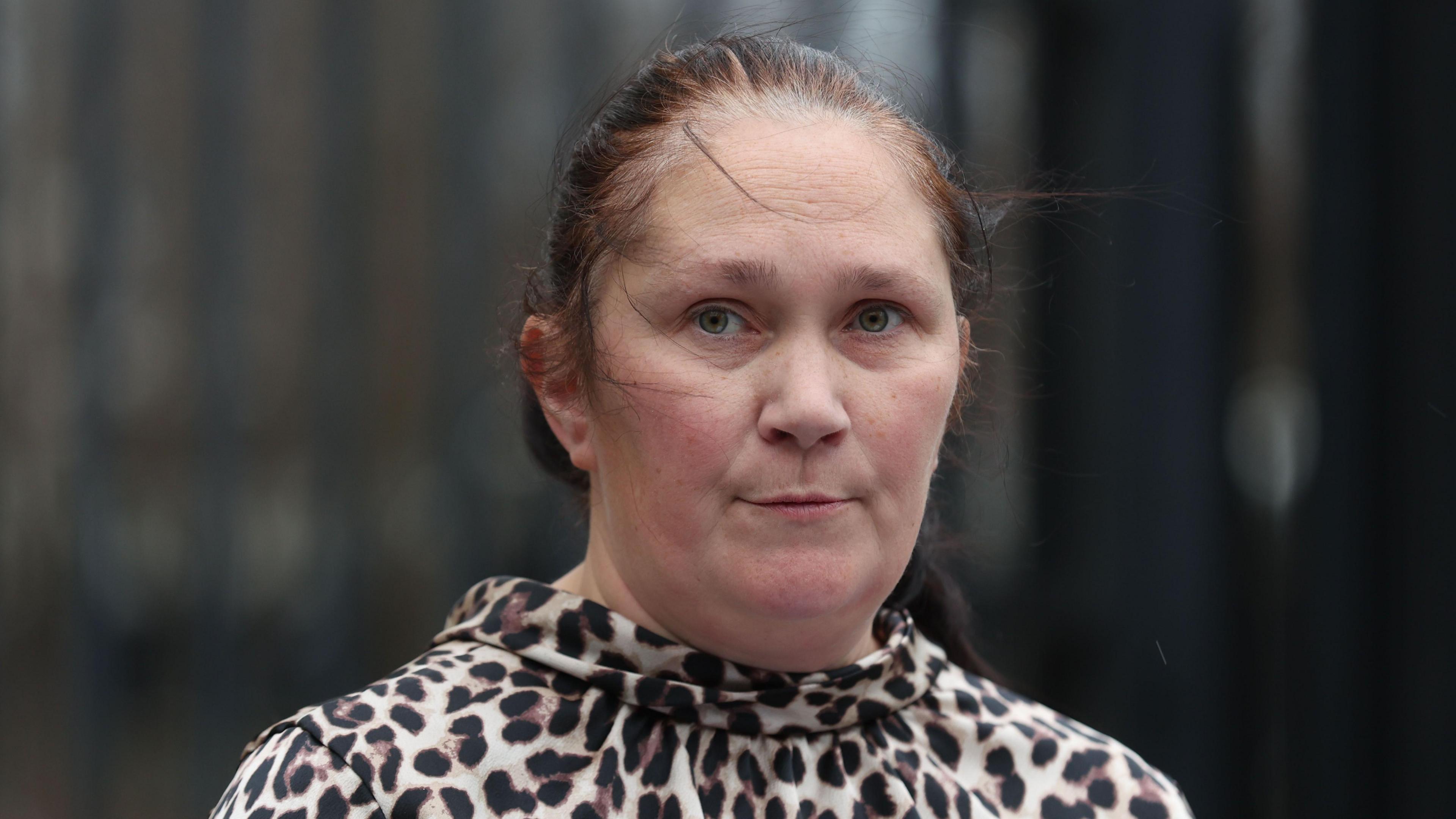 A woman, standing in front of a beige building, looking at the camera. She has brown hair, tied back. She is wearing a leopard print top. 