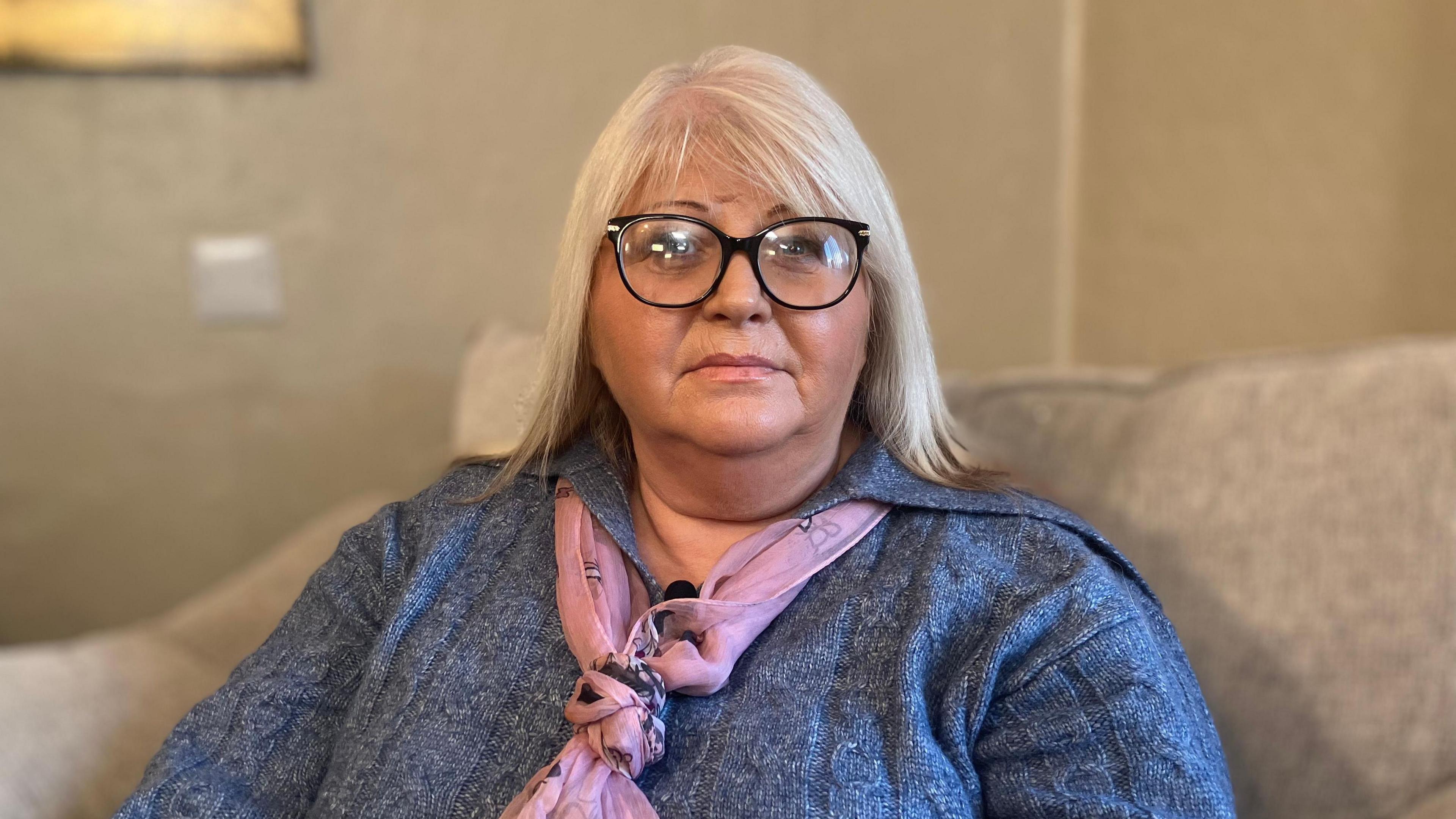 A woman with blonde hair, wearing glasses. She is sat on a a light-coloured sofa. She is wearing a blue top with a pink scarf.