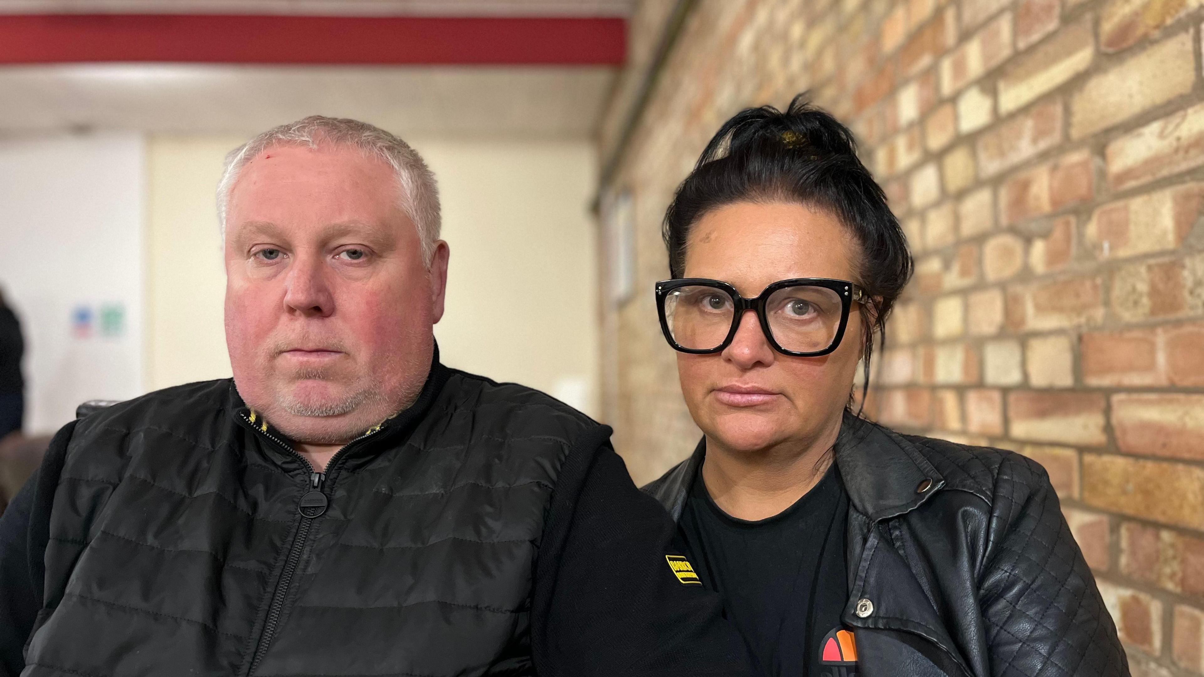 Mark and Caroline Willgoose: a man and a woman both wearing black jackets and staring into the camera. Mark has short white hair, while Caroline has bobbed black hair and is wearing thick-rimmed spectacles. A brick wall is over Caroline's left shoulder.