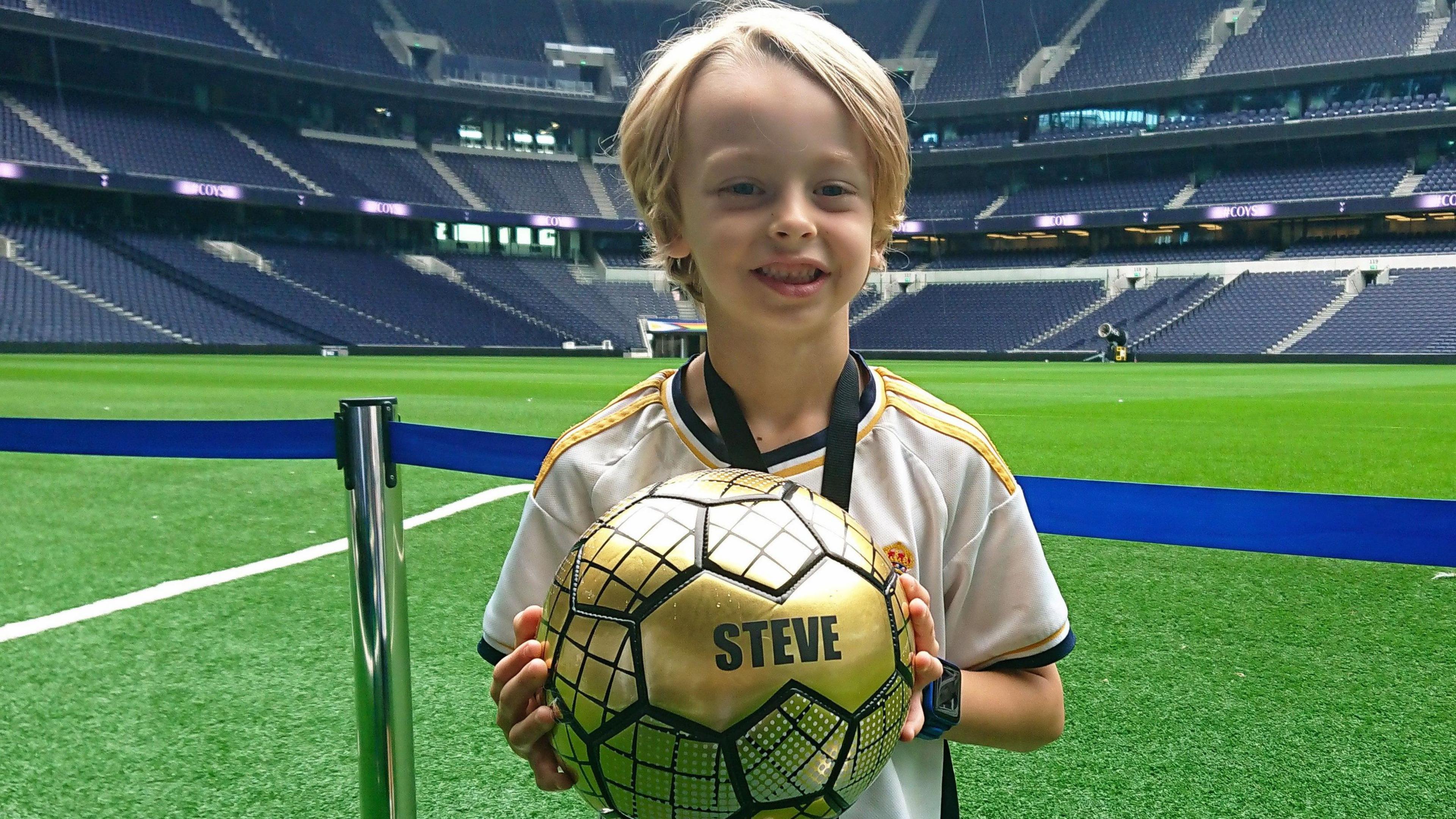 A picture of Zac during a visit to the Tottenham Hotspur Stadium in London. Zac is wearing a football jersey and holding a golden football, specially made in honour of Zac's uncle, Stephen Realf.