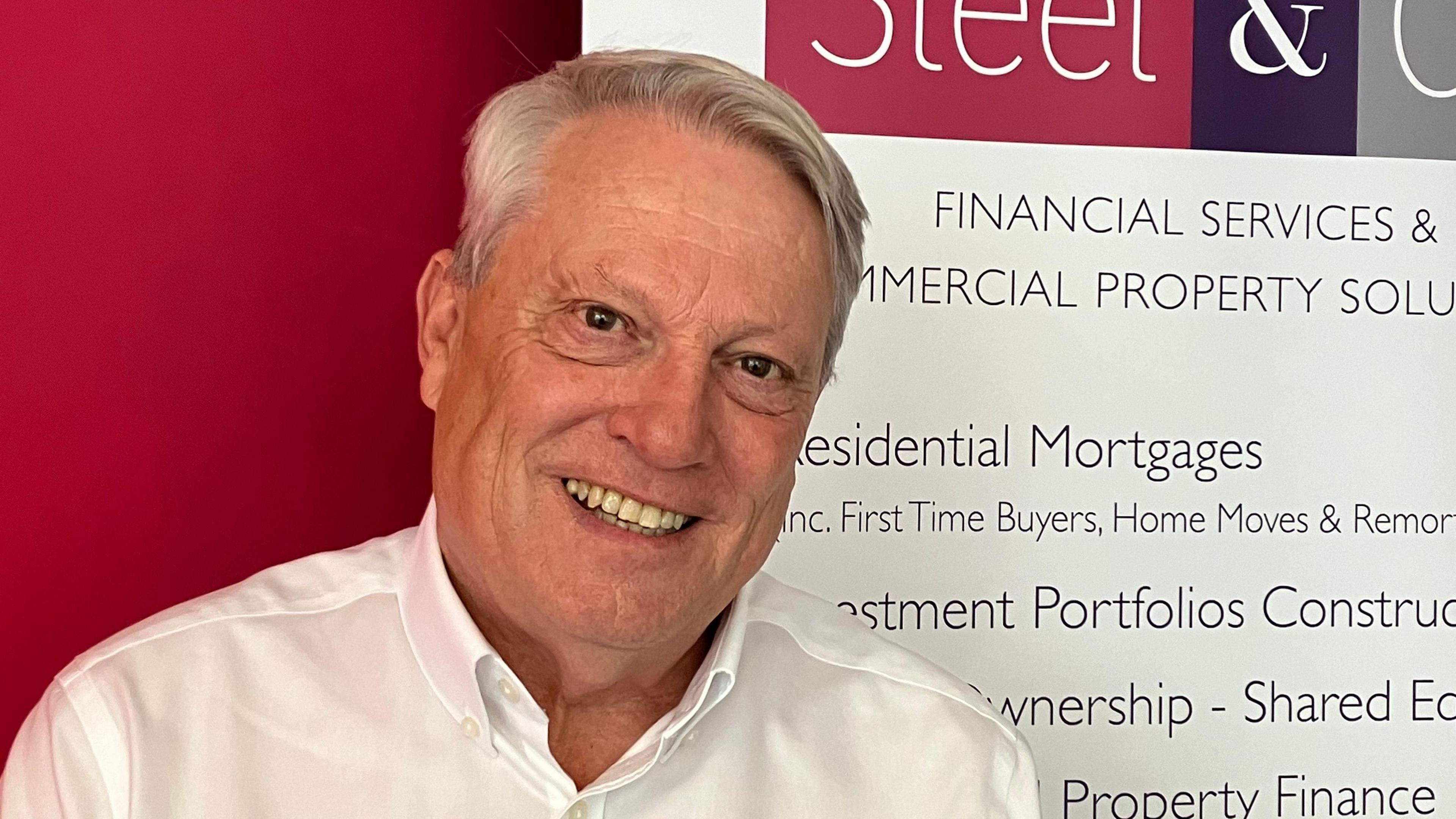 Person wearing white shirt and smiling stands in front of a red and white company board  