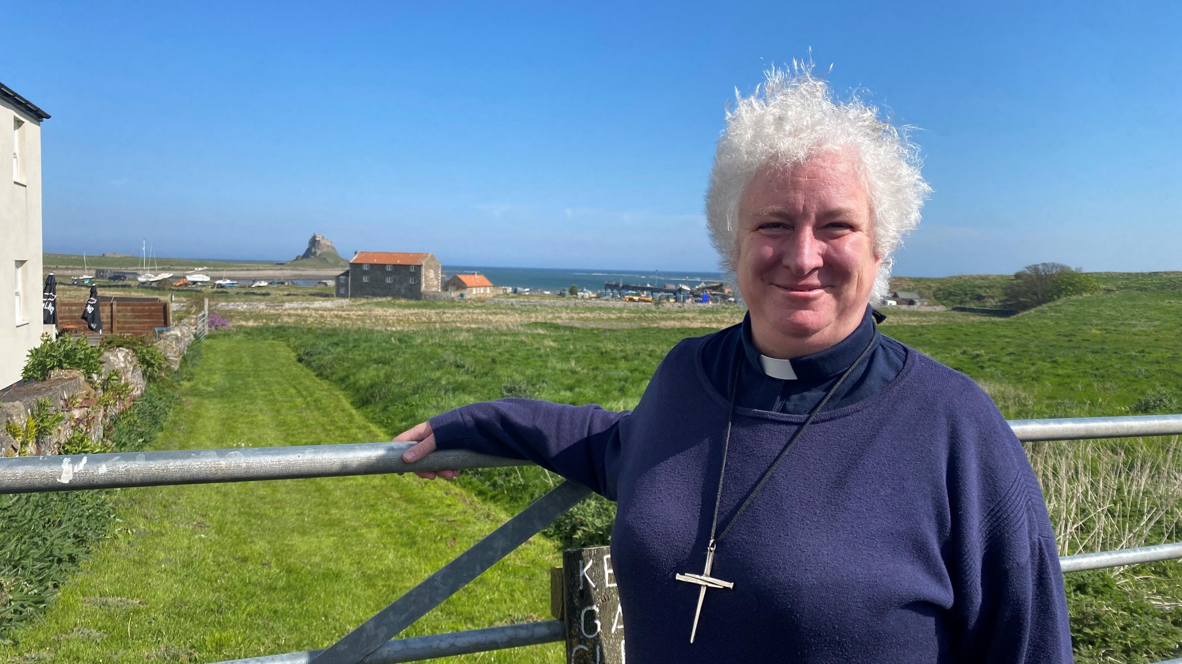 A picture of Sarah Hills, who has short wavy white hair, standing at a gate with the fishing harbour in the far distance behind her and Lindisfarne Castle behind that. She wears a cross around her neck.