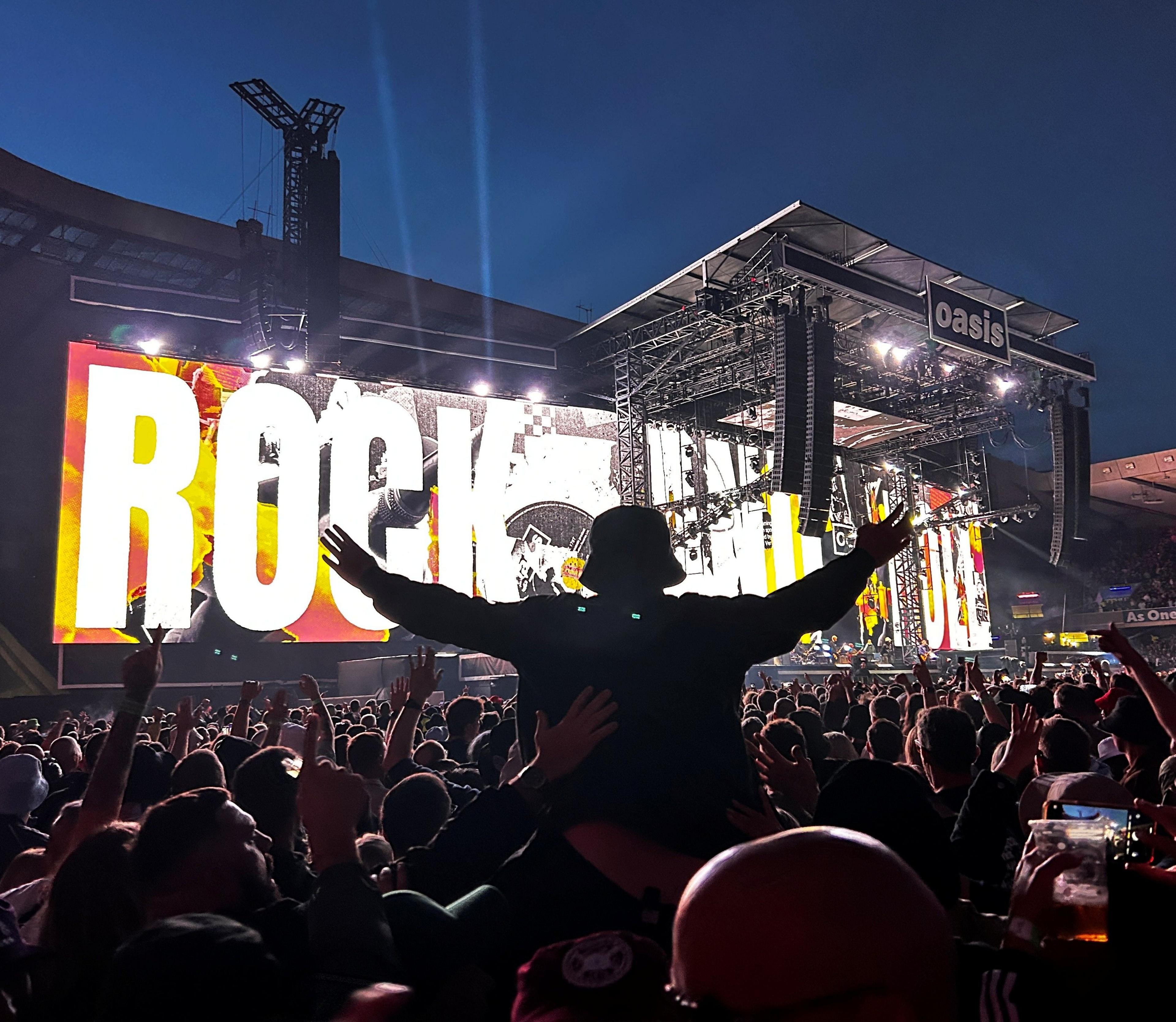 Large crowd at a concert photographed from behind - one person is raised in the air with their arms out, wearing a bucket hat with a large display on stage screens reading "Rock N Roll"