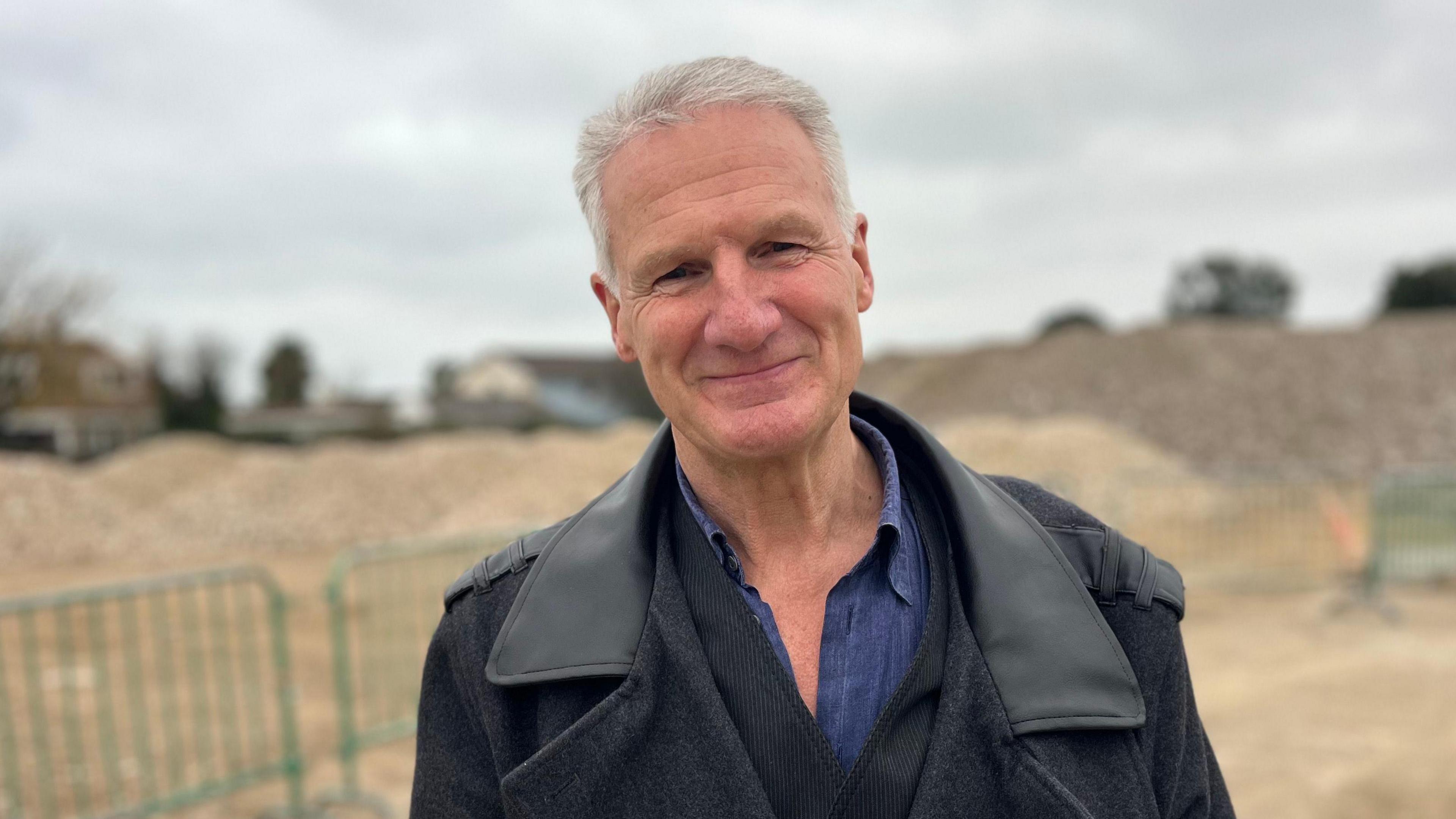 A headshot of Tom Binet. He is smiling directly at the camera and is wearing a dark grey coat and a blue shirt. He has short grey hair. He appears to be standing near a building site.