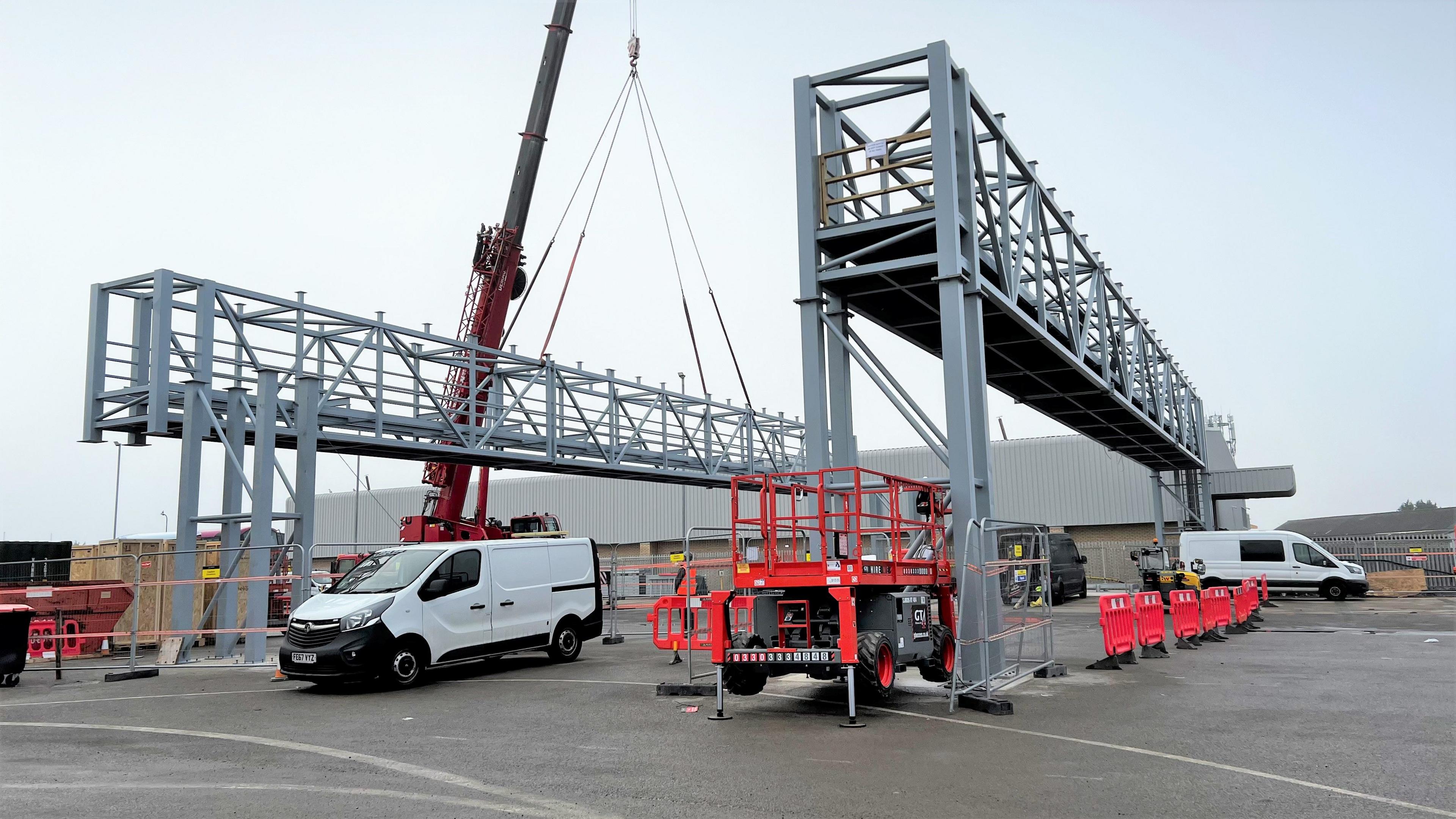 A red crane lifting a huge metal gantry into place to recharge electric buses at the Weston bus depot. There are white vans parked on the site and red barriers preventing anyone from walking underneath the gantries.