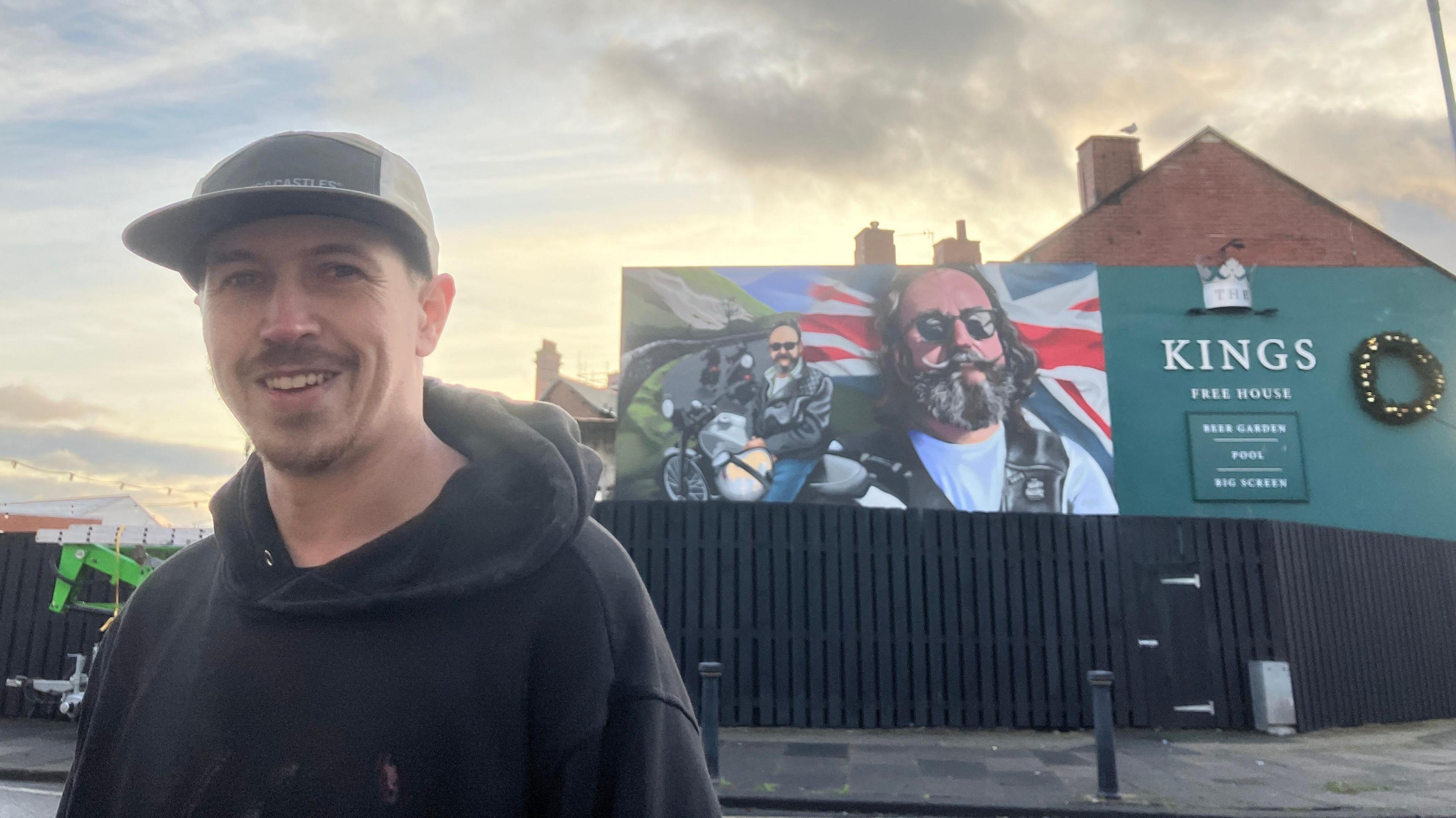 Artist Shawn Sharpe stands in front of the mural he has created in Barrow. It shows two images of Dave Myers - one sitting on a motorbike and another of his head and shoulders. Behind him is a mountain scene and a union flag. It is painted on the side of a pub called Kings Free House.