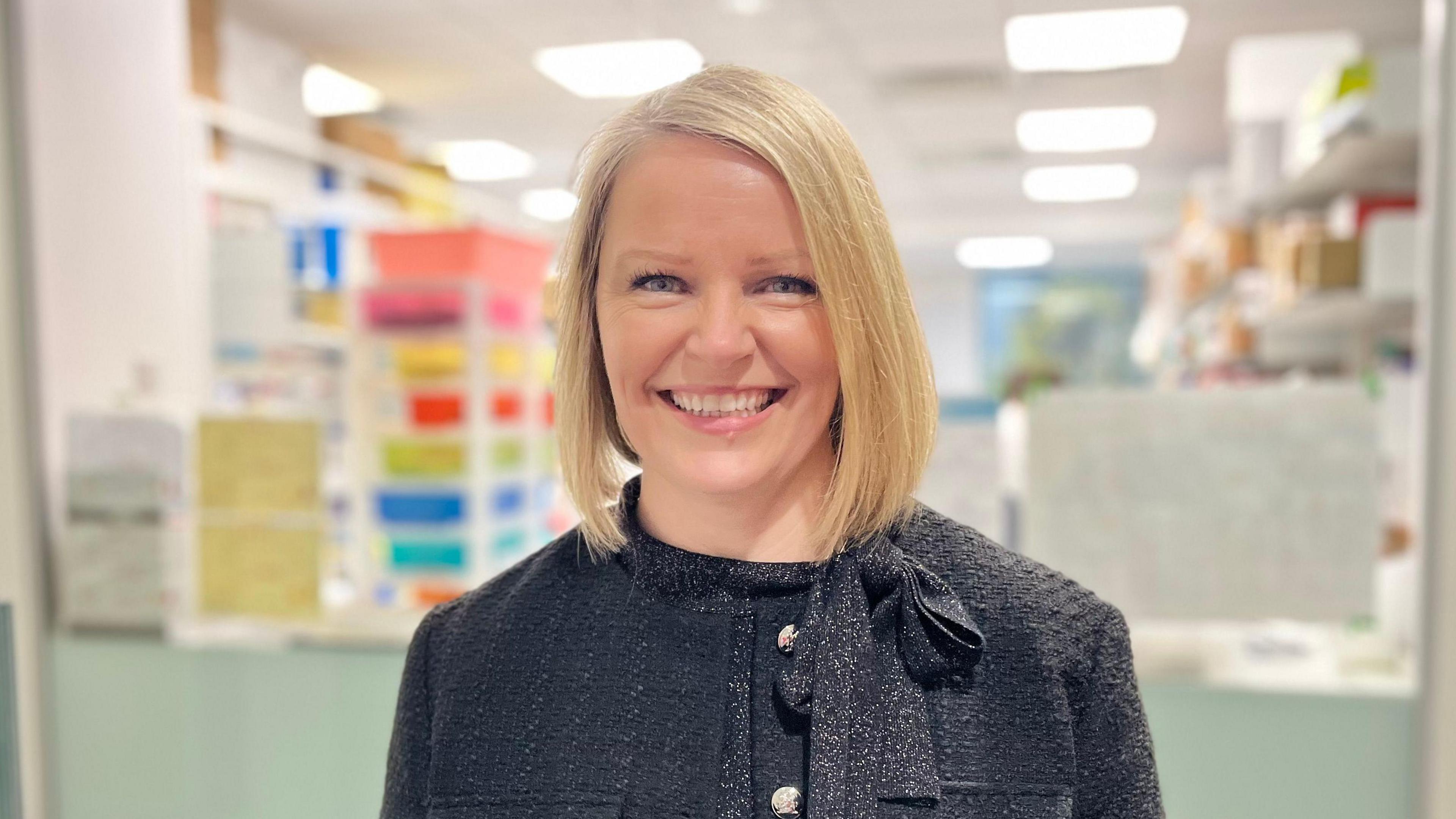 A woman with blond hair, wearing a grey jacket smiling at the camera. She is stood in a pharmacy setting 