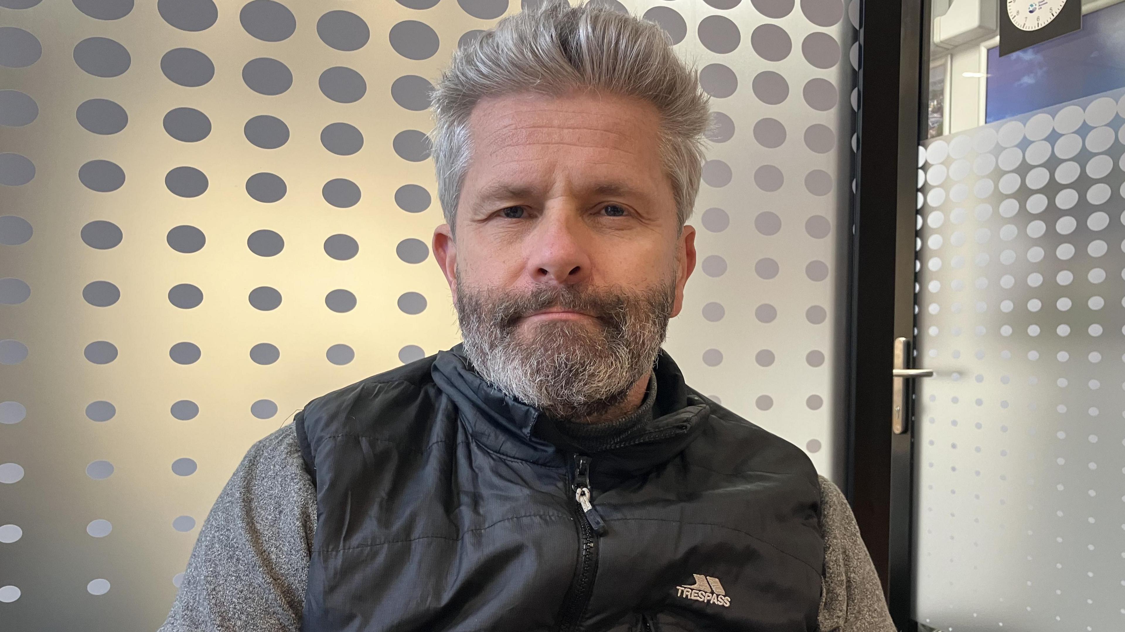 A man with silver hair and a black body warmer, in front of a window.