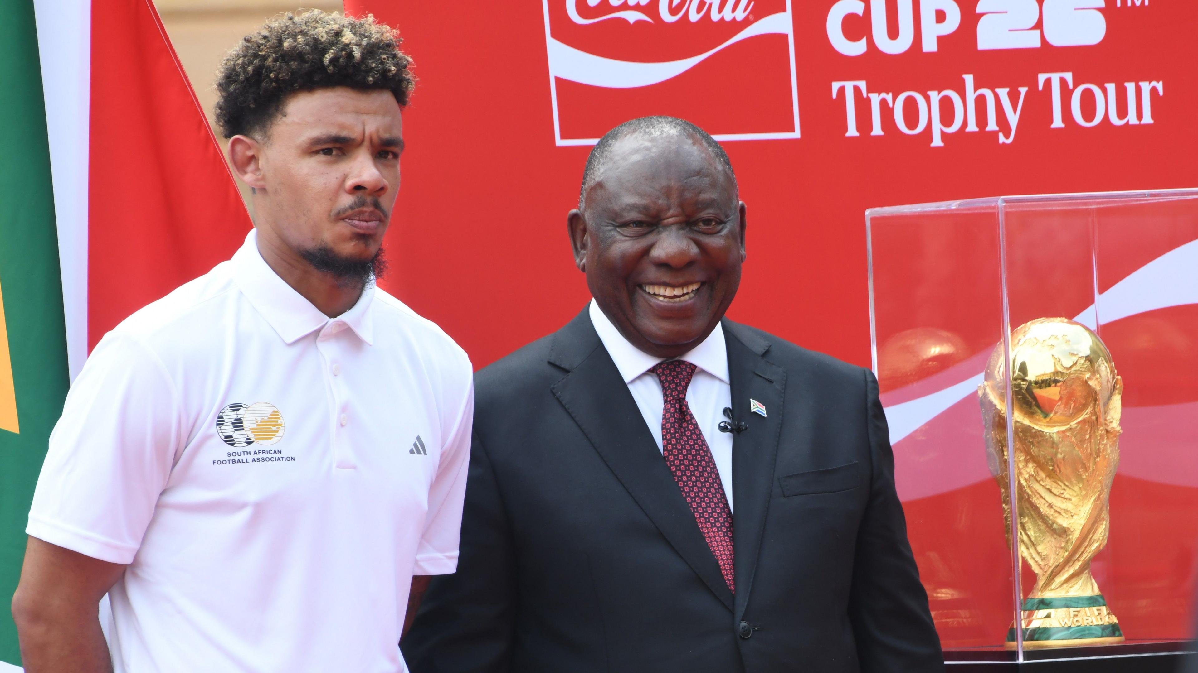 Ronwen Williams and South Africa's President Cyril Ramaphosa stand alongside each other with the World Cup trophy in a clear box next to them. Williams, on the left of the image, wears a stern expression and a white polo shirt with the emblem of the South African Football Association, while Ramaphosa wears a dark suit, white shirt, red tie and a beaming smile