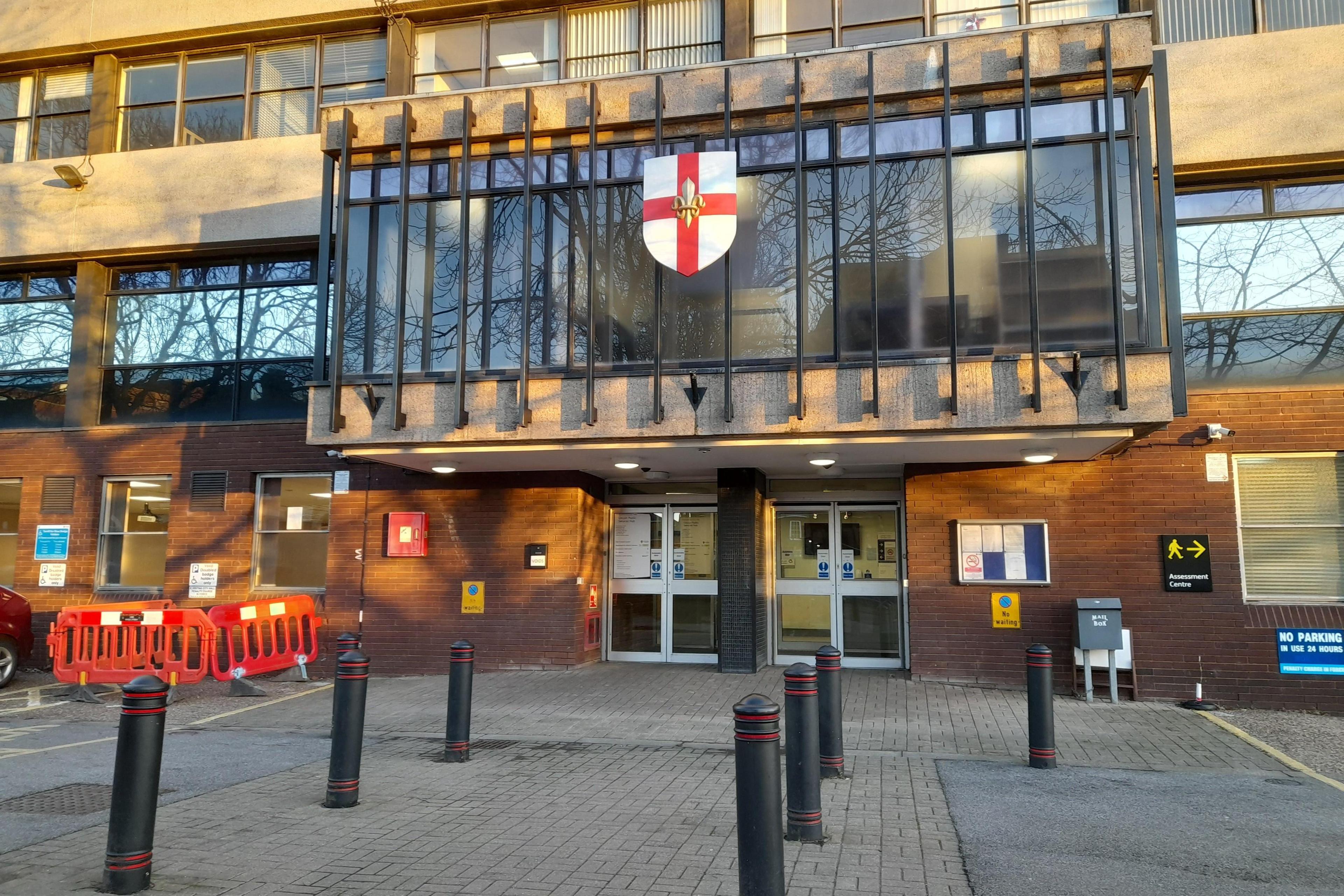 The front of a large mid-20th Century public building. Three stories are in the picture with rows of windows separated by smooth cladding. There are two sets of double doors and above them a large feature window with black horizontal bars and a shield motif decorated with the cross of St George.