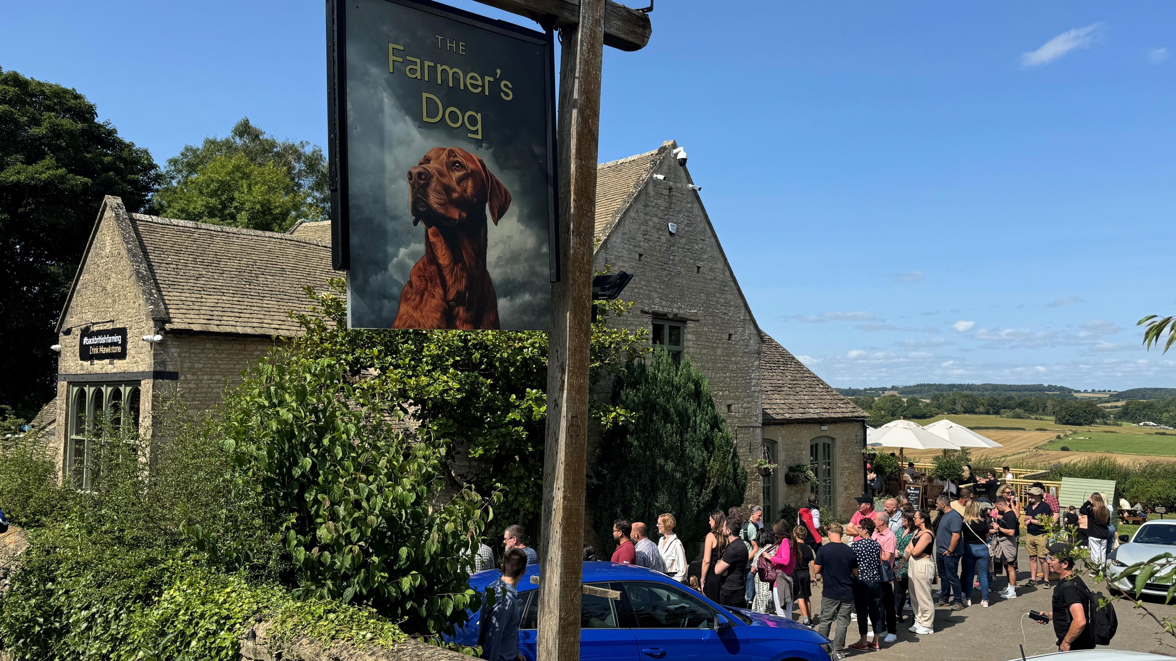 An historic pub under blue skies with rolling fields in the distance and a pub sign with a dog in the foreground