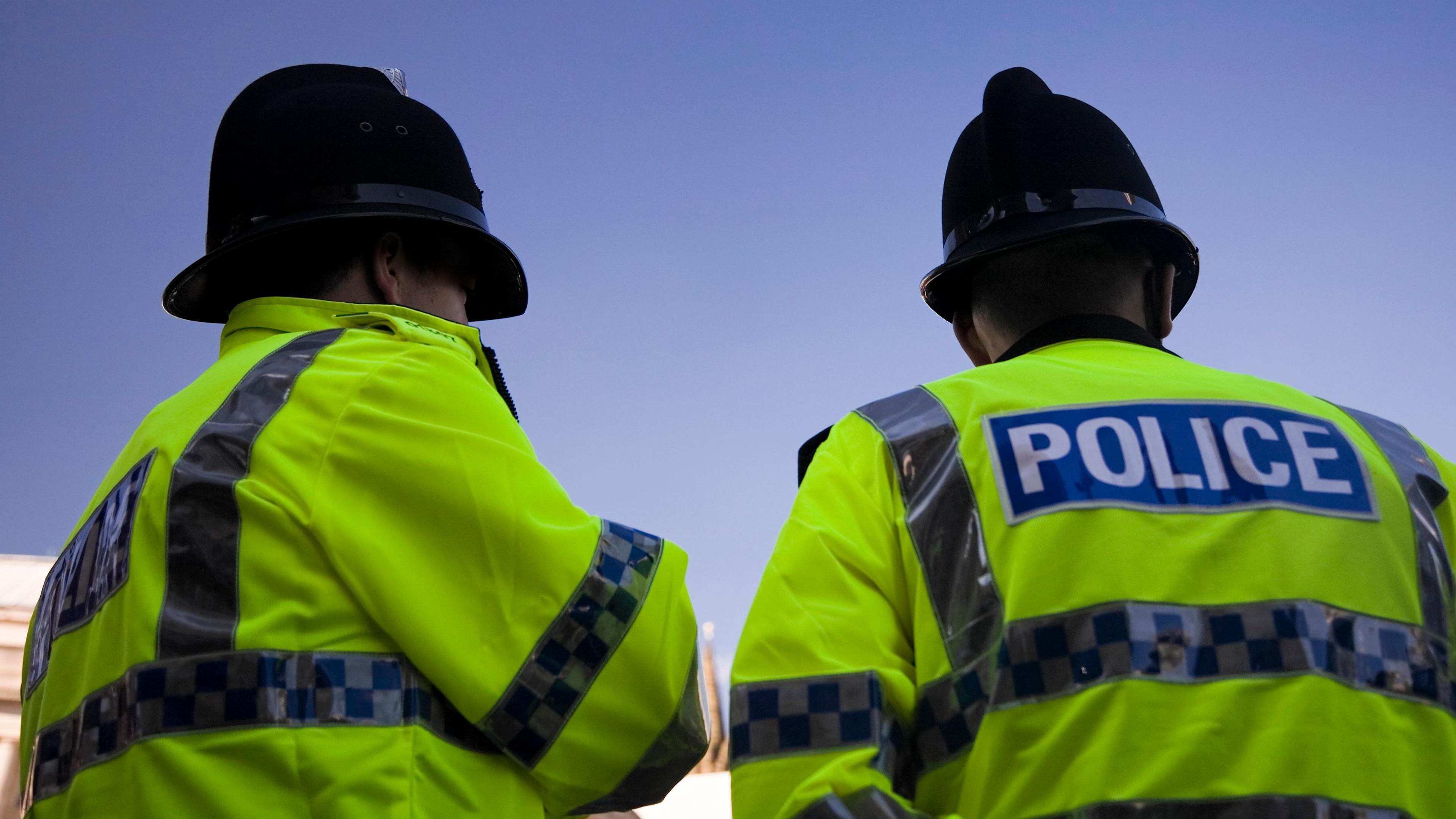 The backs of two police officers wearing police jackets and helmets, looking towards a blue sky.