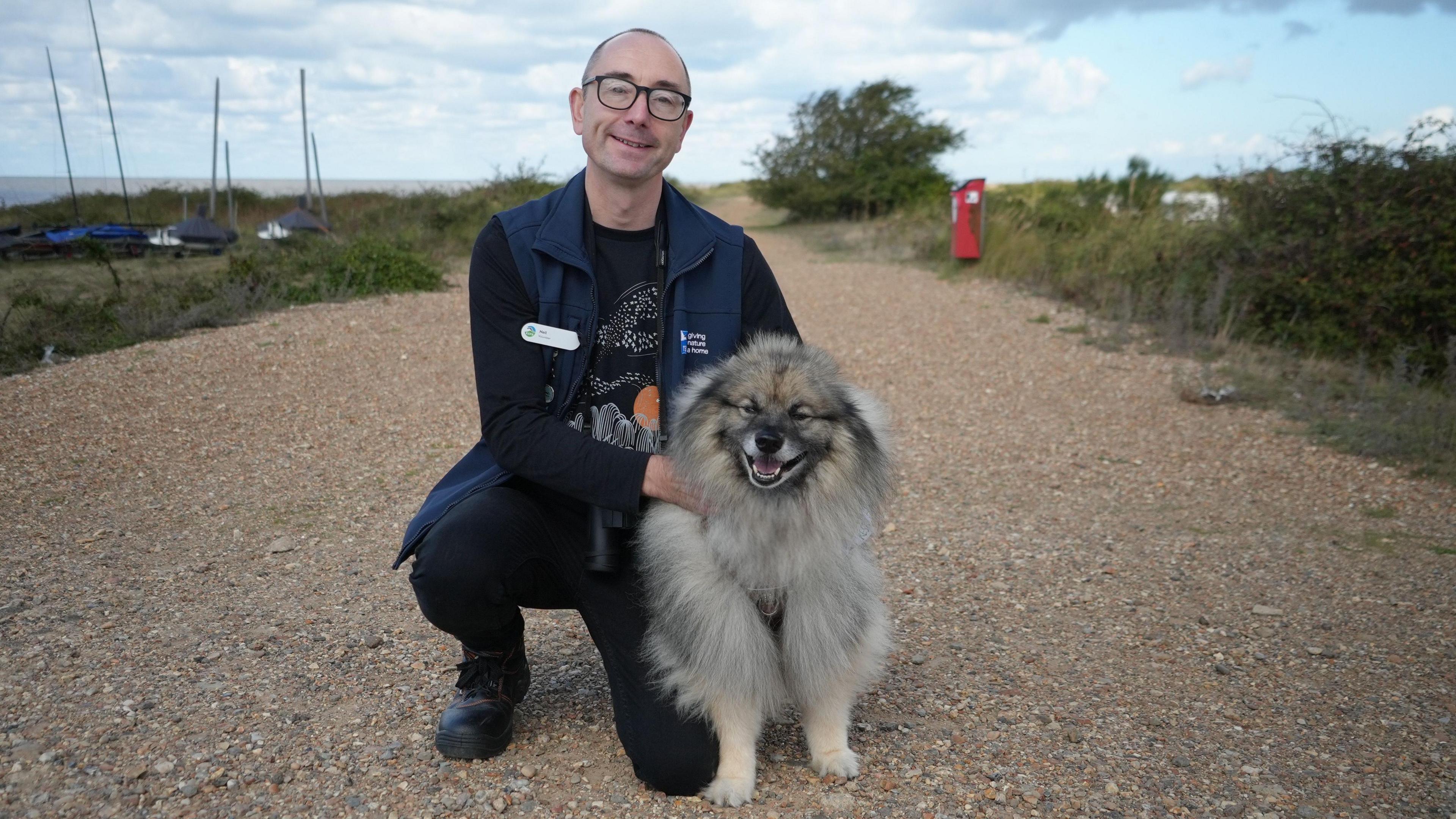 Man with bald head and glasses and dark jacket with a fluffy grey and brown medium-sized dog with cream-coloured legs.