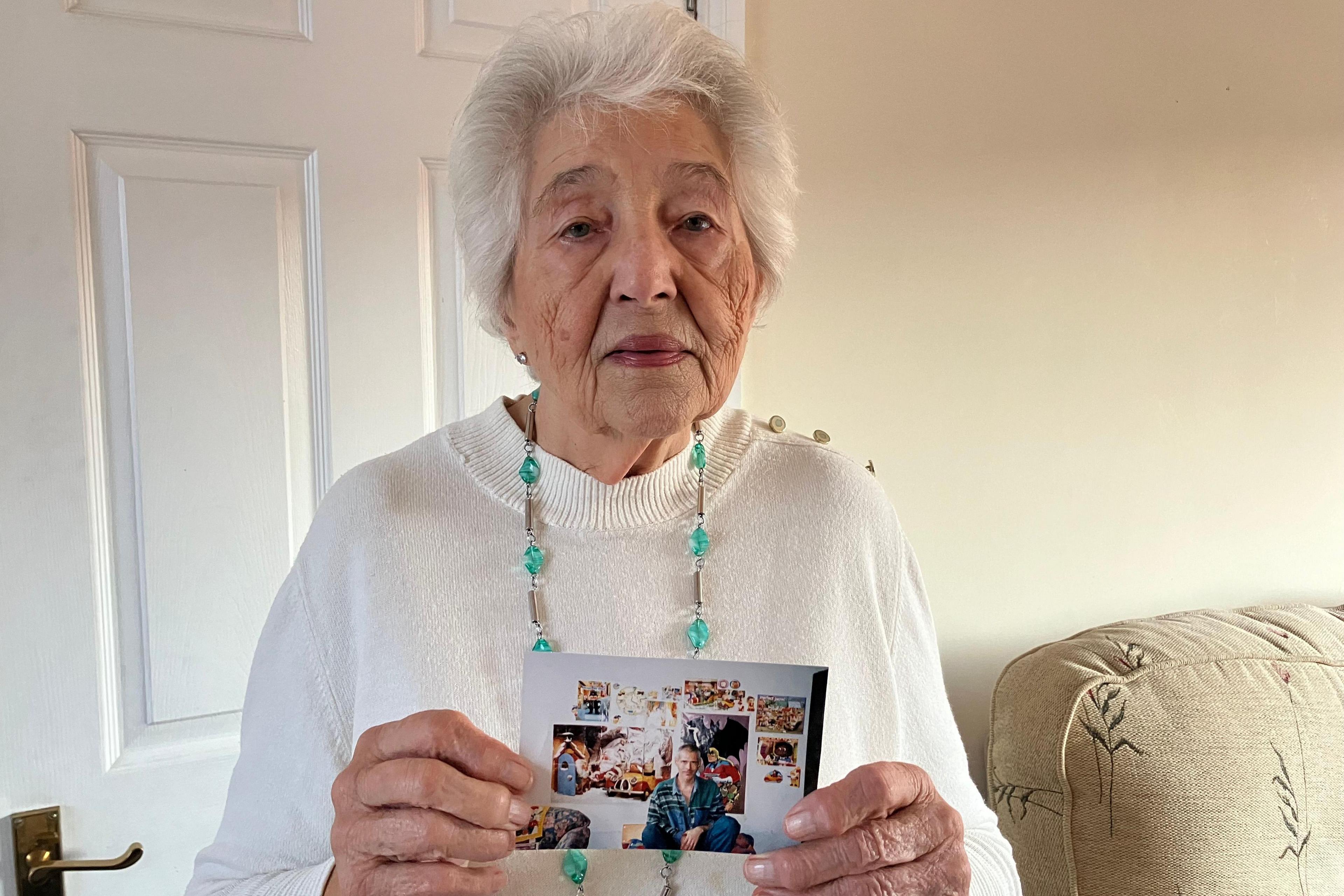 A head and shoulders picture of Heather Pratten, holding up a photograph of her son Nigel. She is in a living room, wearing a white jumper. She has white hair and is wearing a long emerald neckless.