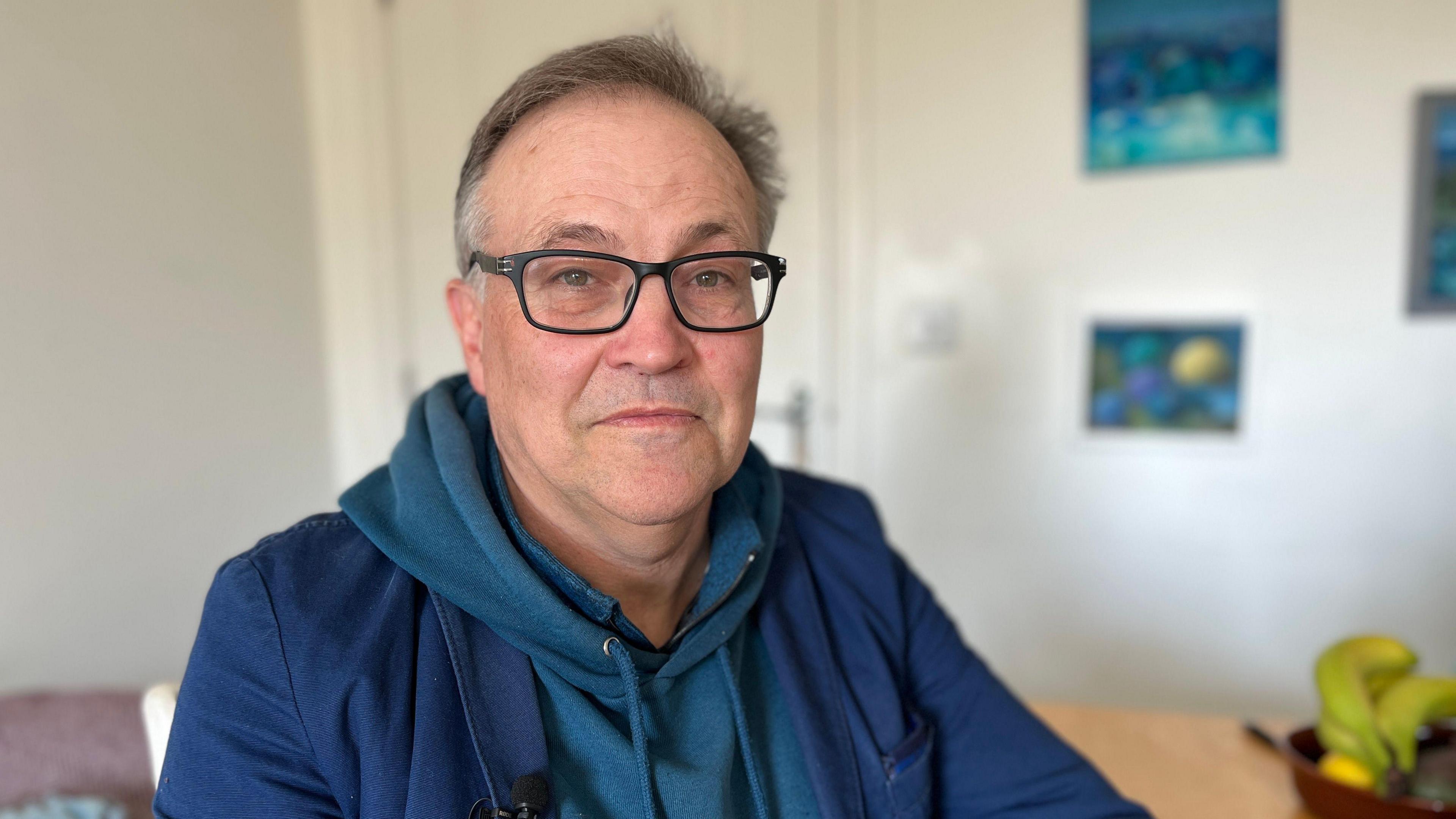 A head and shoulder shot of Philip, who has grey hair and wears a pair of spectacles. He is wearing a blue hoodie with a blue cardigan on top of it. In the background you can see various paintings and some bananas in a basket.