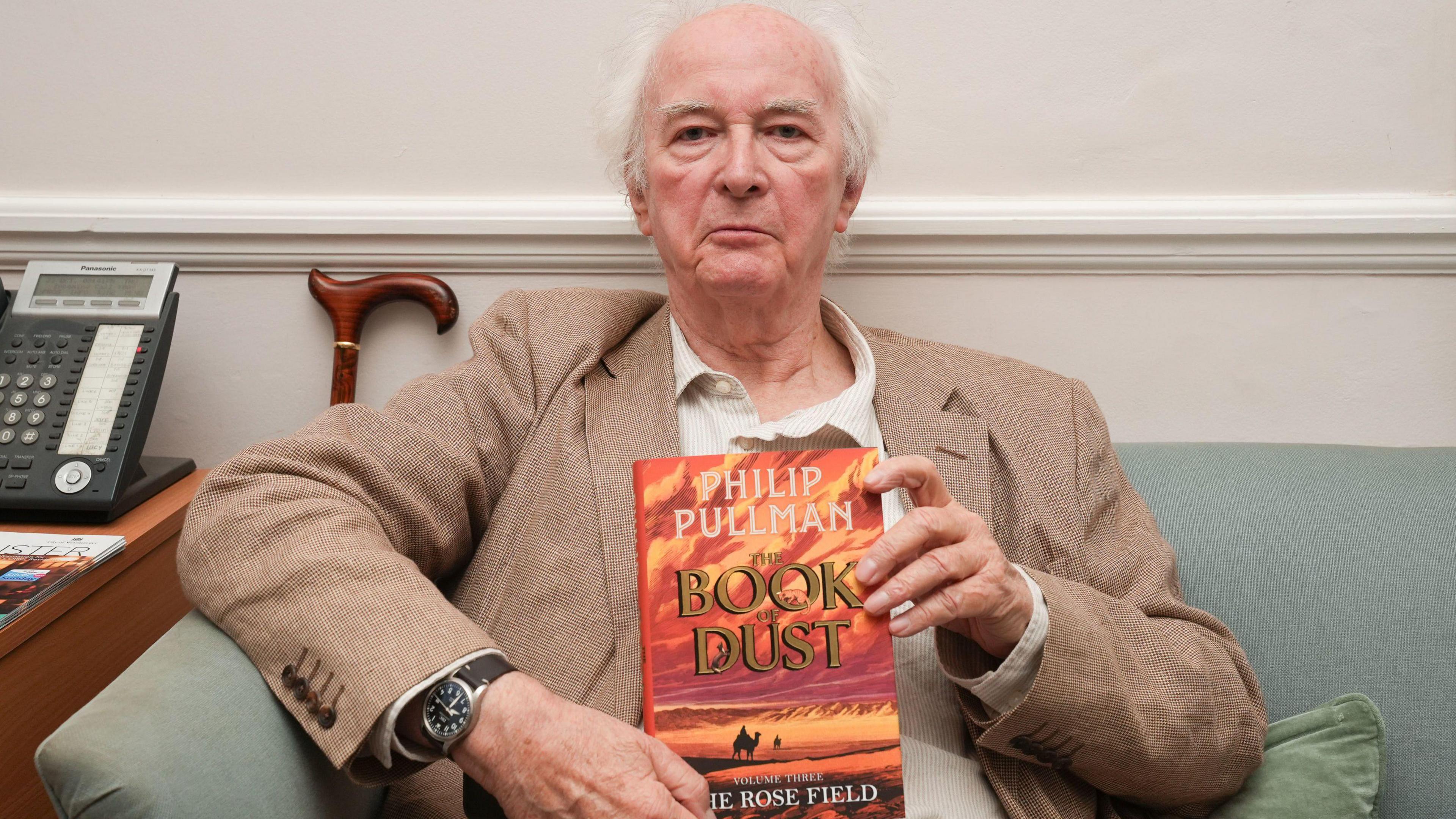 Philip Pullman at the launch event for the long-awaited conclusion to his bestselling The Book of Dust and His Dark Materials sequence: The Rose Field at St James's Church, Piccadilly in London. He is sitting on a sofa and holding his book. There is a cane behind him and a telephone on a table next to him.