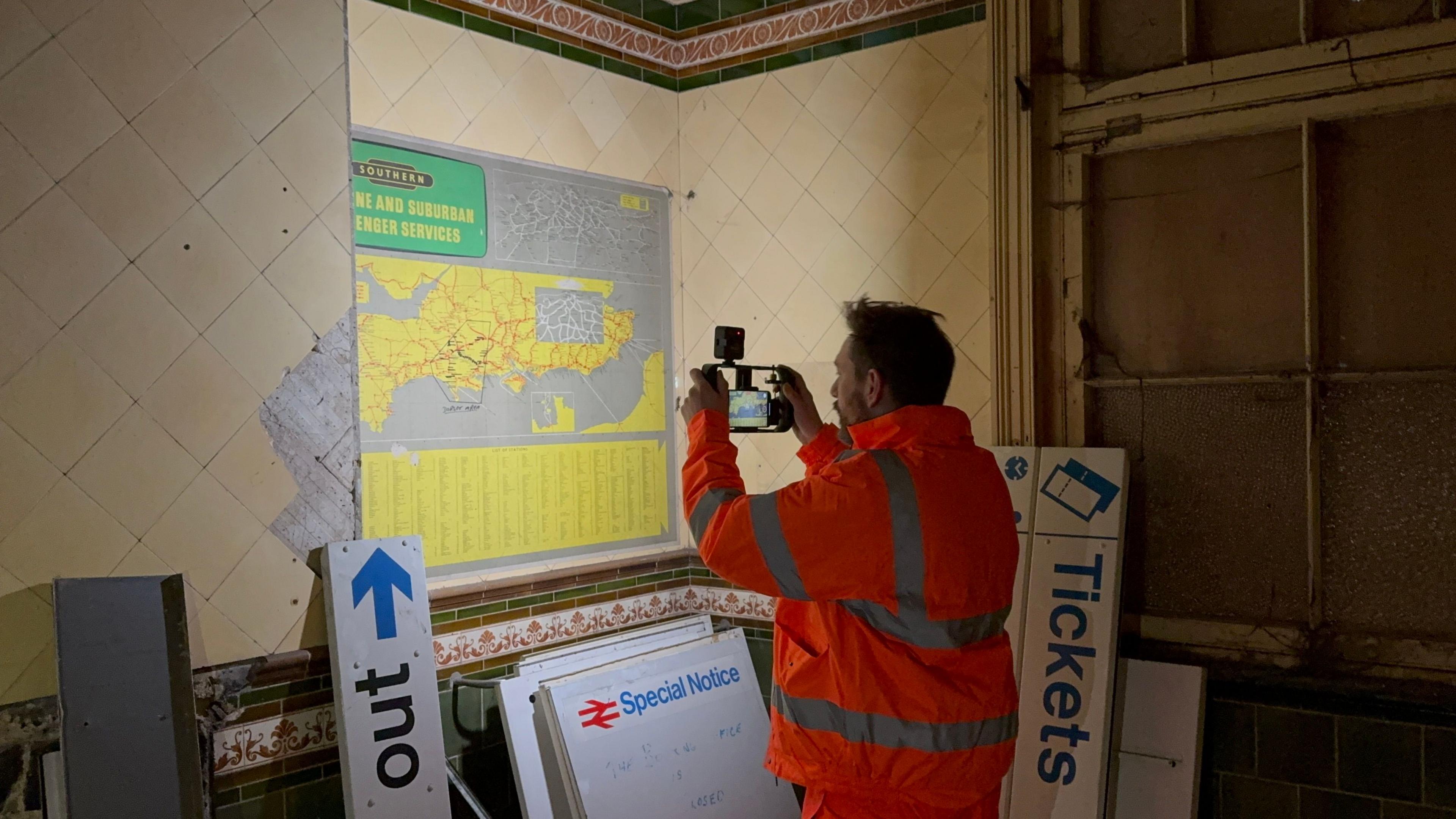 A man wearing an orange hi-vis suit and holding up a phone and a torch to take a picture of a map on the wall of an underground room. The walls are covered in cracked and yellowing tiles, and there are noticeboards and ticket signs stacked up in piles. The grey and yellow map is outdated.