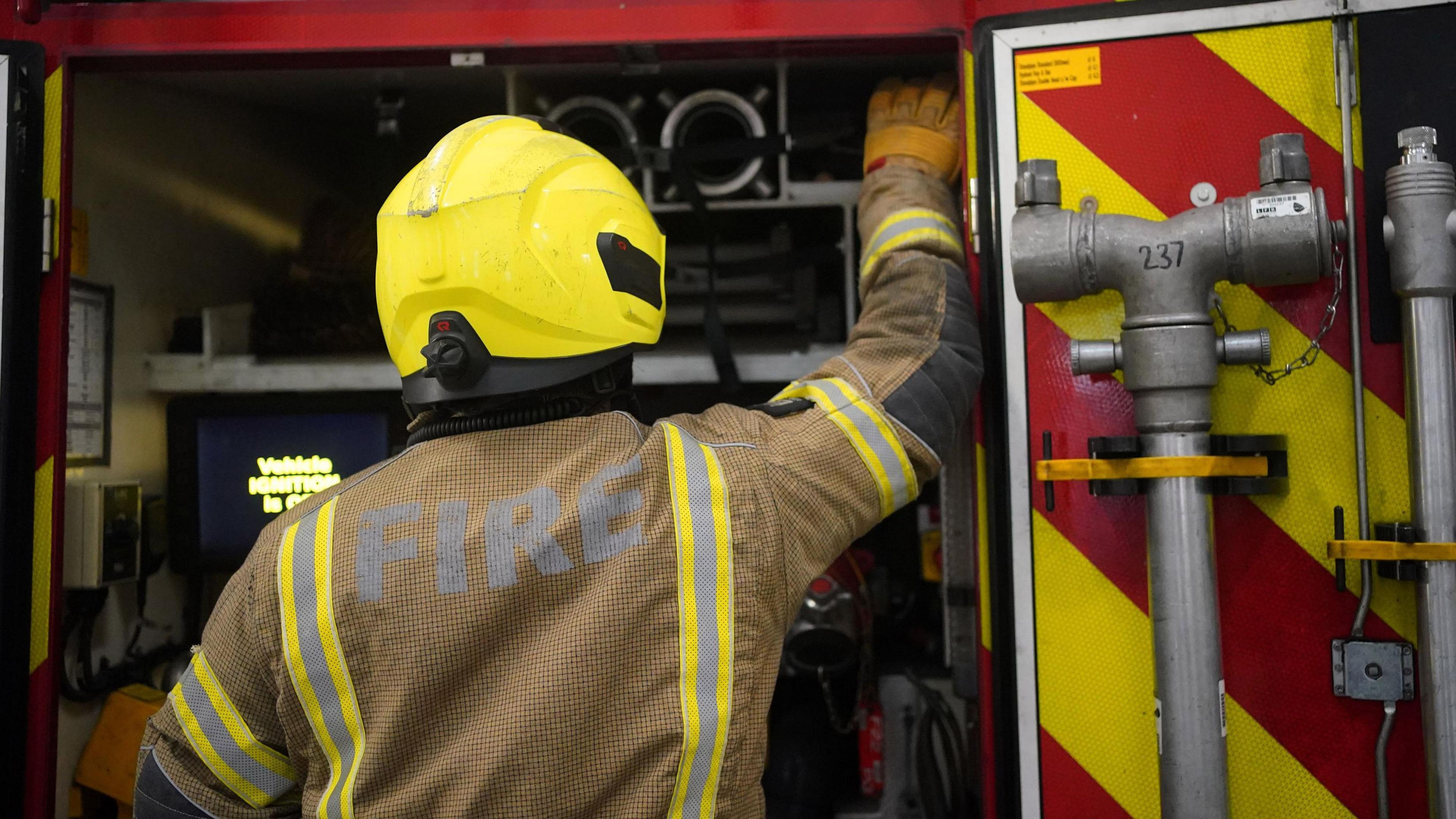 A firefighter in uniform and yellow helmet is standing facing the rear end of a fire engine. The firefighter is removing equipment from the red and yellow truck. Pieces of heavy equipment are hanging on the door.