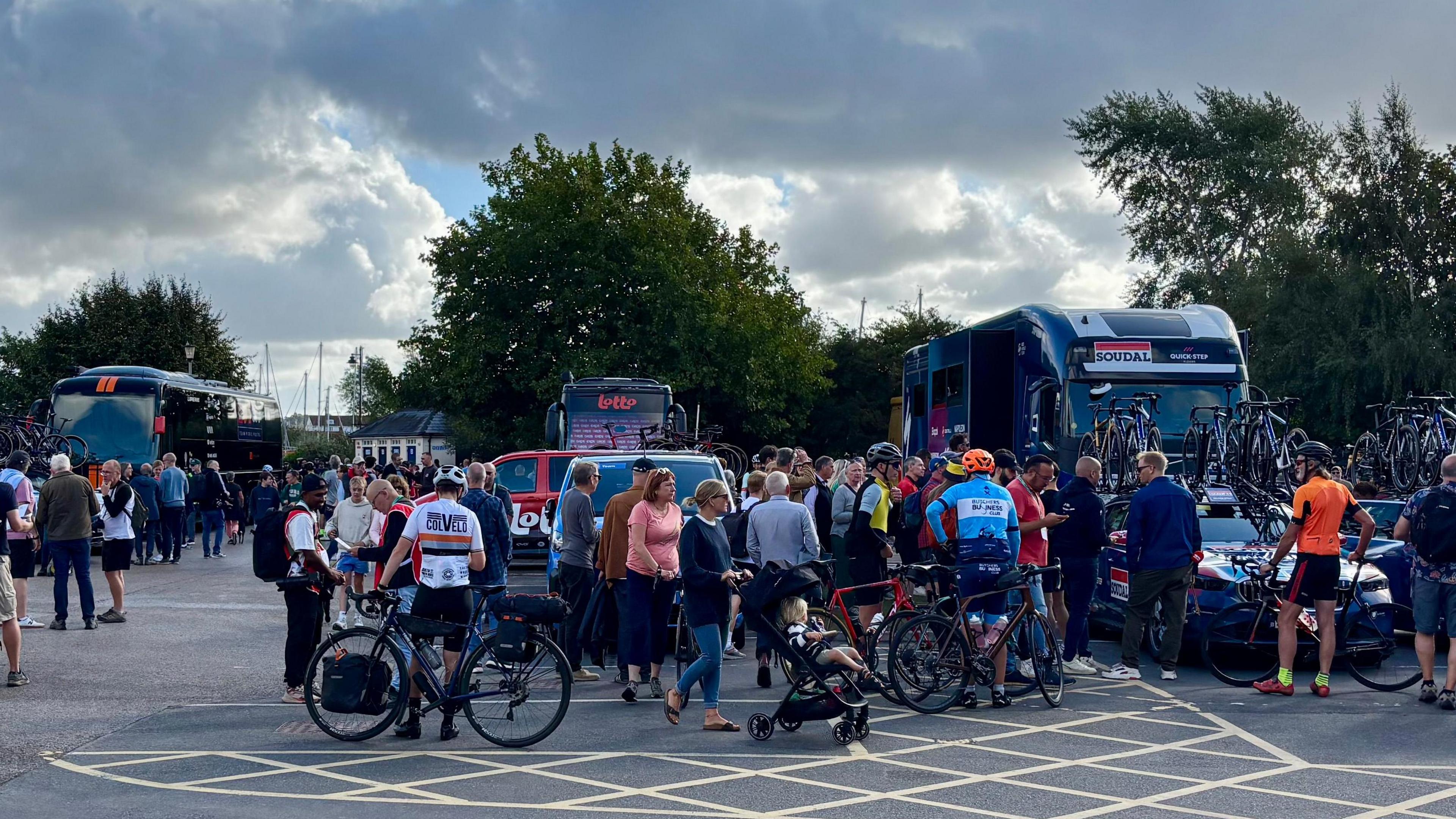 Groups of people stand in a car park. There are some riders stood with their bikes as other non-riders talk with them. Team buses can be seen parked up.