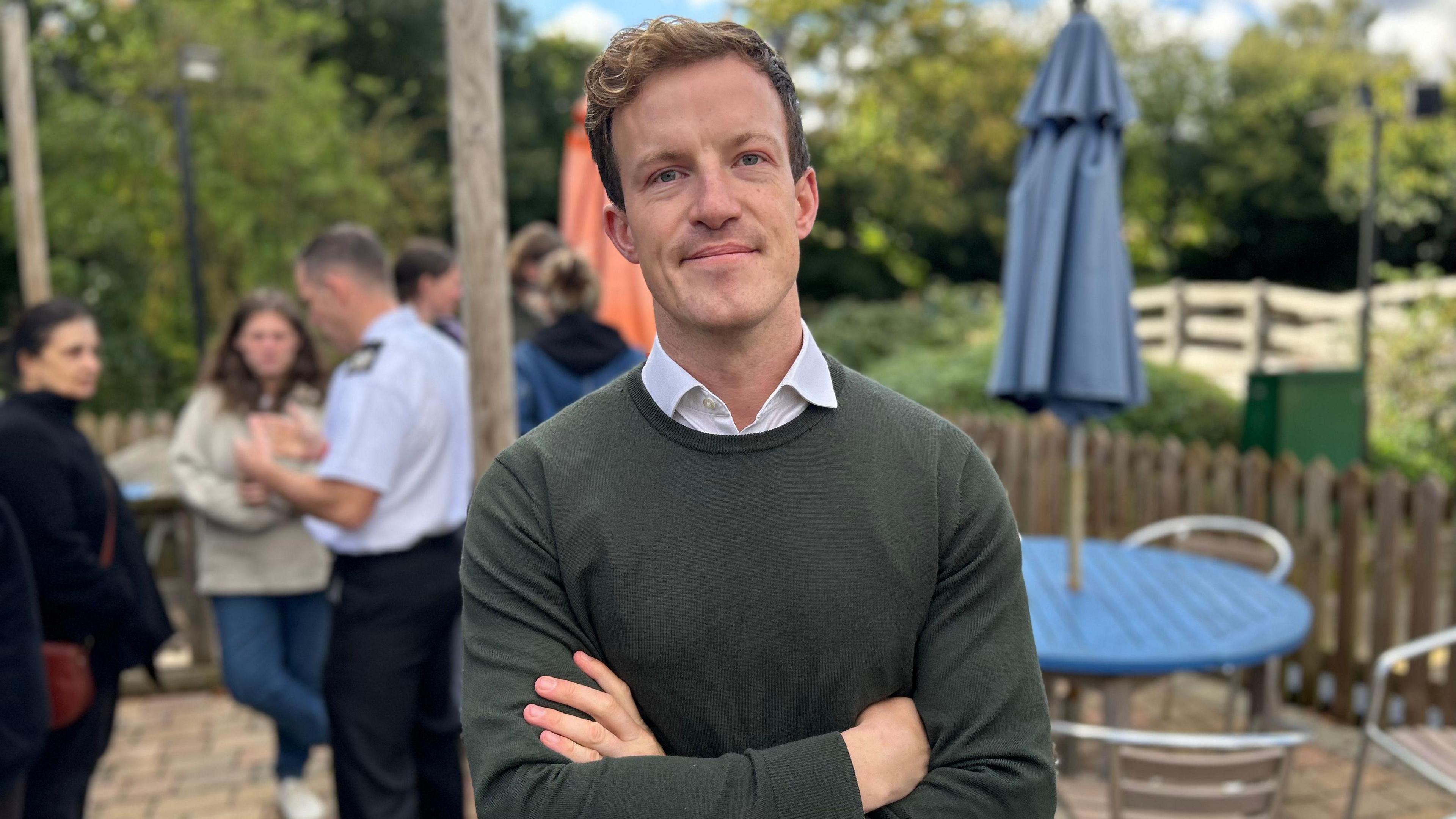 A picture of the Labour MP for Hitchin, Alistair Strathern. He has his arms folded and is wearing a dark green jumper, which is over the top of a white shirt. Faded out in the background are a group of people.