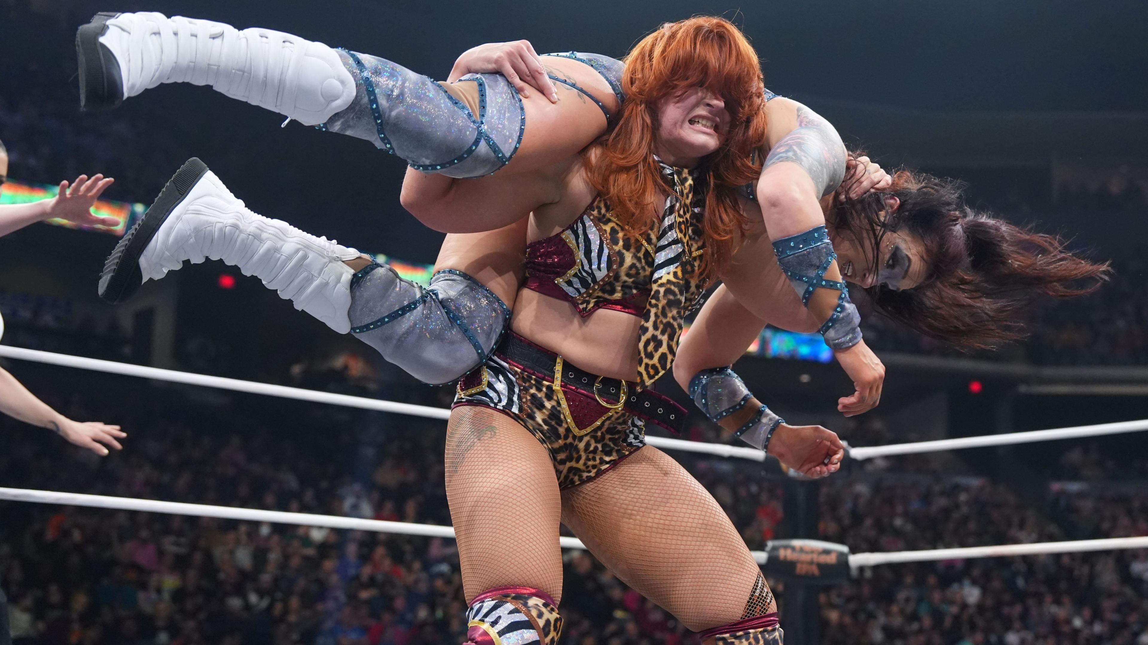 Jamie Hayter - a woman with red hair, wearing a red cropped top and red and gold shorts, holding an opponent on her shoulders in a wrestling ring