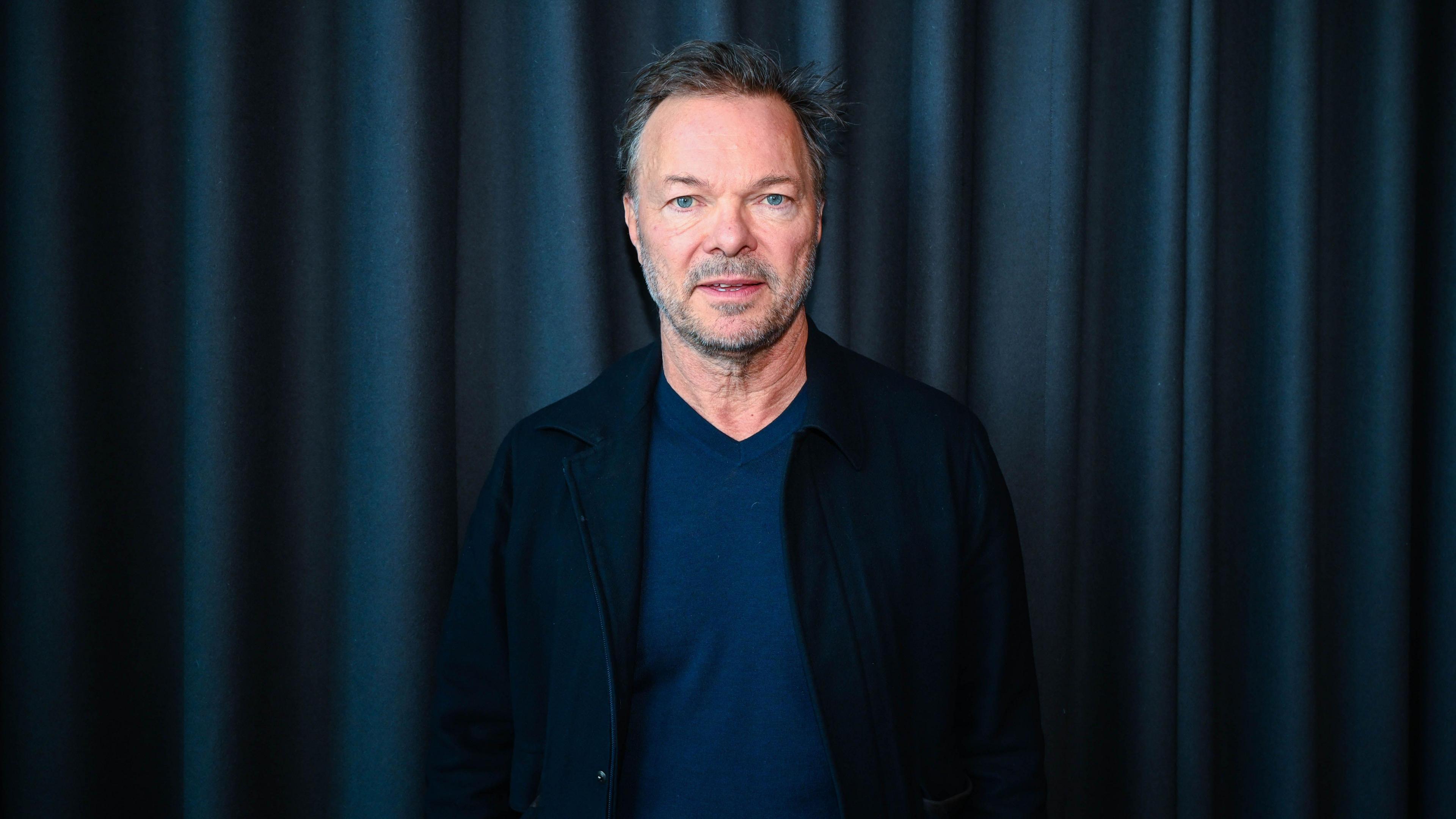 A man (Pete Tong) with short brown hair pushed slightly back has short grey stubble and a moustache. He is looking directly into the camera with his mouth slightly open and has blue eyes. He is wearing a black jacket blazer and blue top with a navy curtain behind him.