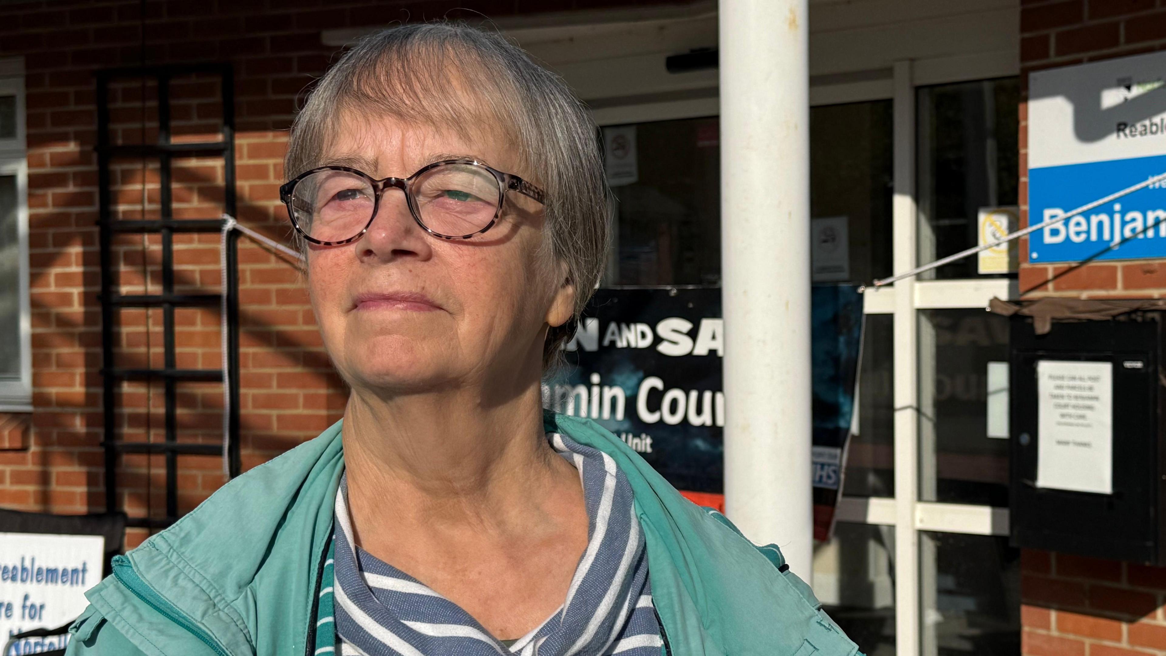 Siri Taylor; a woman who is standing in front of a closed NHS building which was previously Benjamin Court. She is looking away from the camera and is wearing a green coat over the top of a blue and white striped T-shirt.