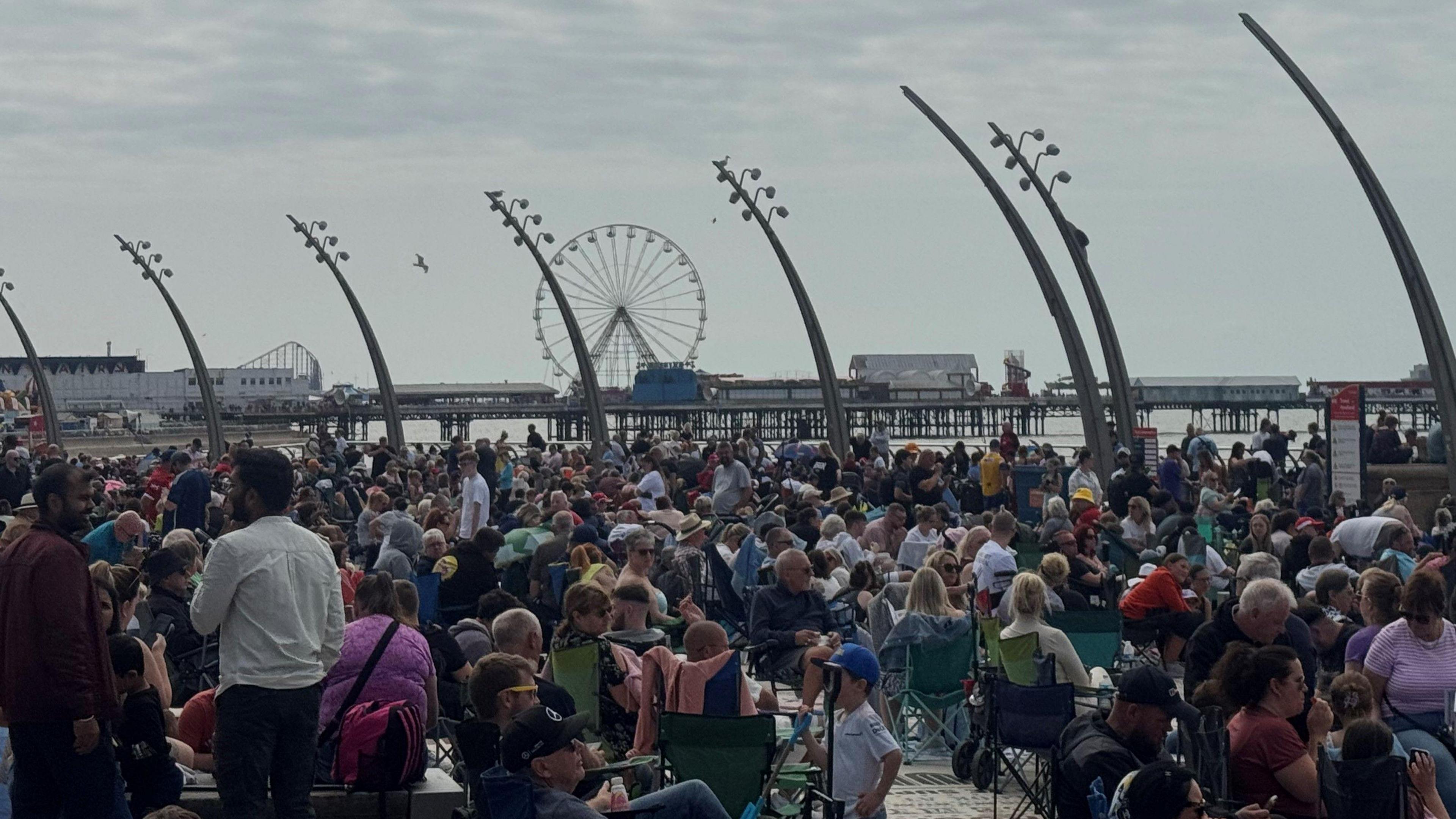 Hundreds of people sit on the promenade with a pier jutting out to sea in the background. A large ferris wheel and a rollercoaster can be seen in the backdrop.