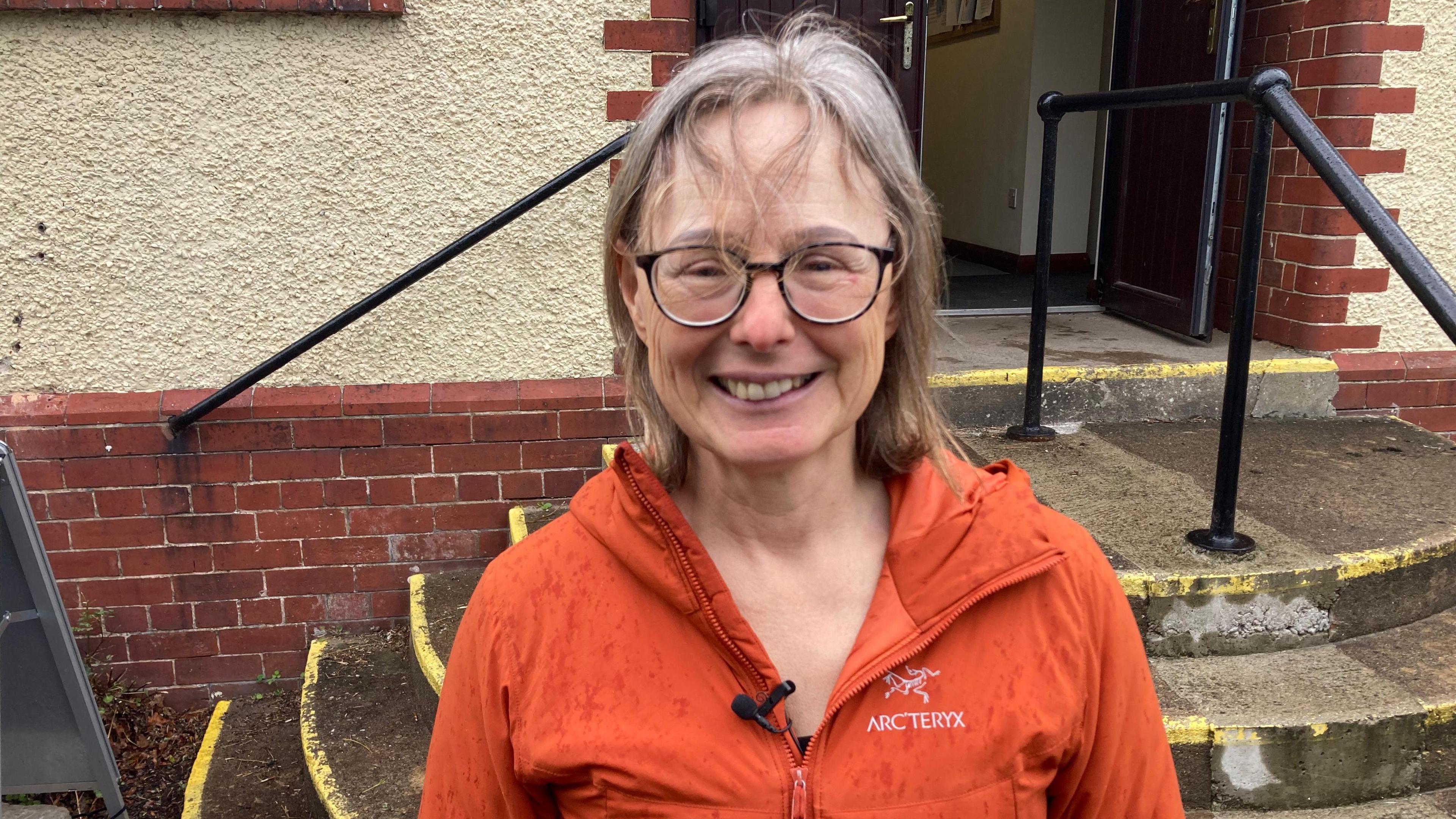 Pindar is standing close to the camera, facing forward. She is wearing a bright orange waterproof jacket with a visible front zip and a small clip-on microphone attached near the chest area. The jacket appears to be lightly speckled with water droplets, reinforcing the impression of wet weather. The jacket also shows a printed brand name, “ARC'TERYX” on the upper chest.