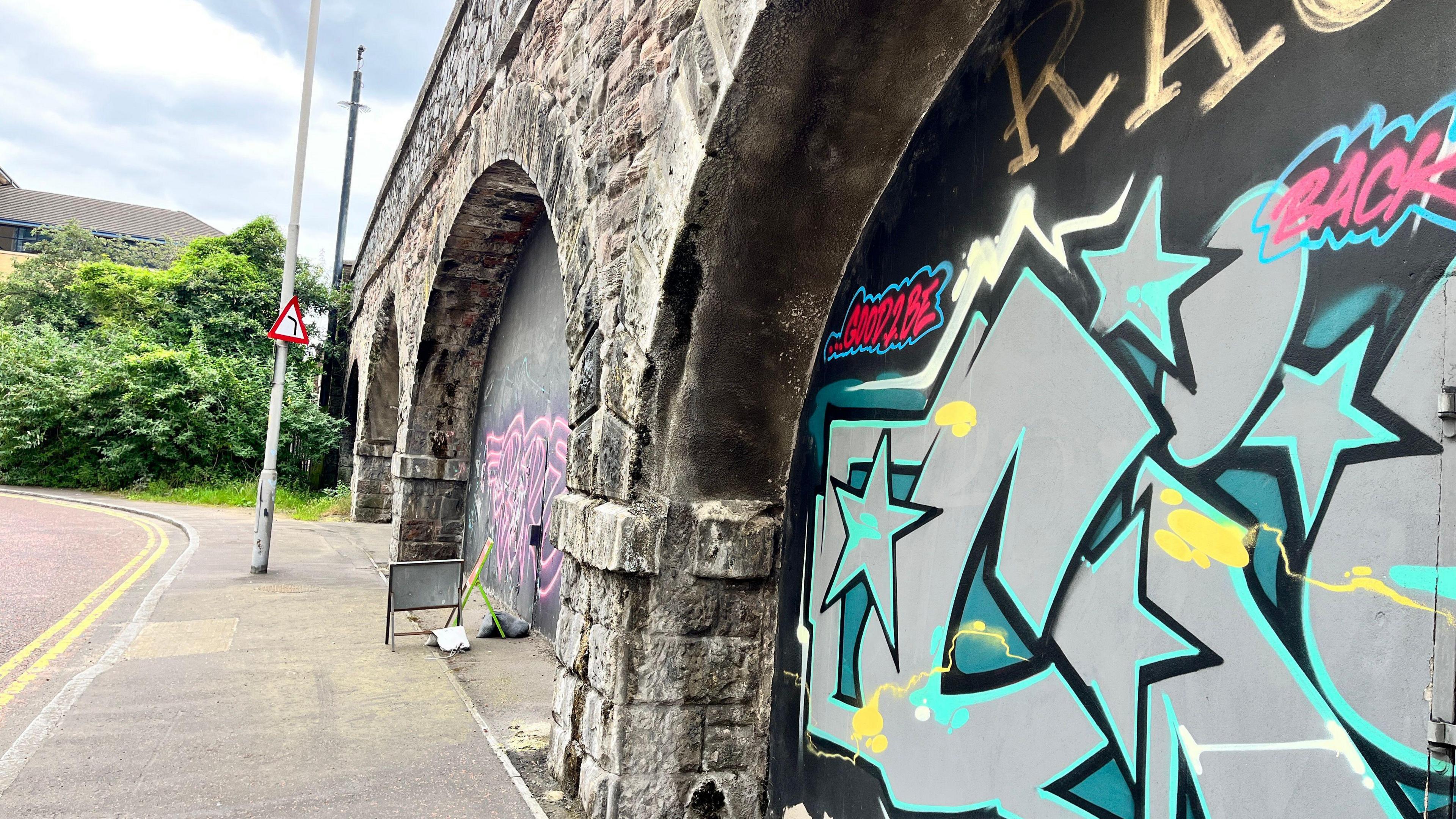 A grey stone bridge with graphited walls inside of its arches. There is a footpath running alongside the bridge and double yellow lines. In the background is a green hedge and single lamp post. Behind the hedge is a large building.