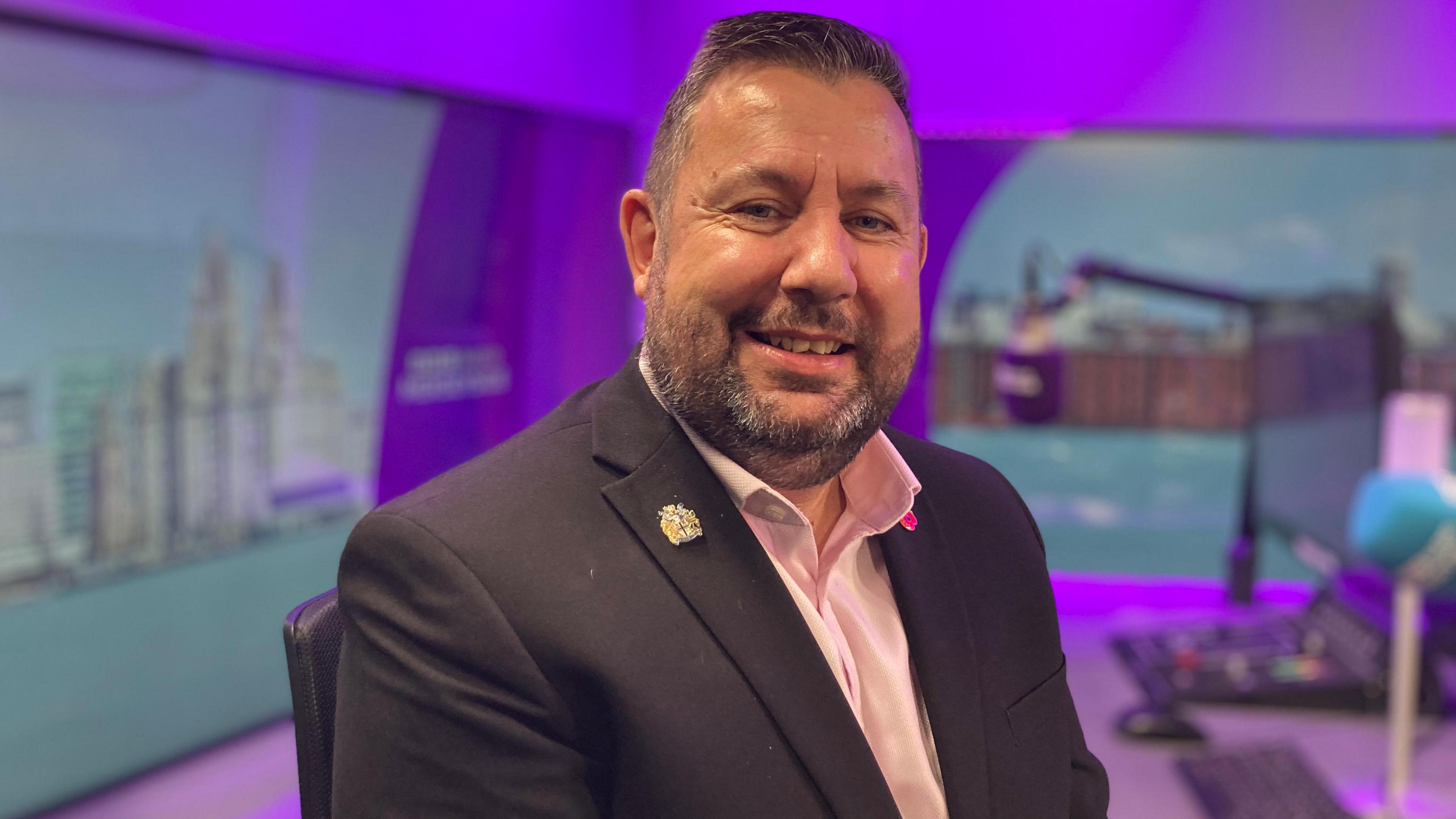 Anthony Burns sits in a radio studio, a blue microphone and control desk are visible in the background. He smiles at the camera. He has short dark hair and a stubbly beard. He wears a dark suit jacket with a pink, open-collar shirt underneath.