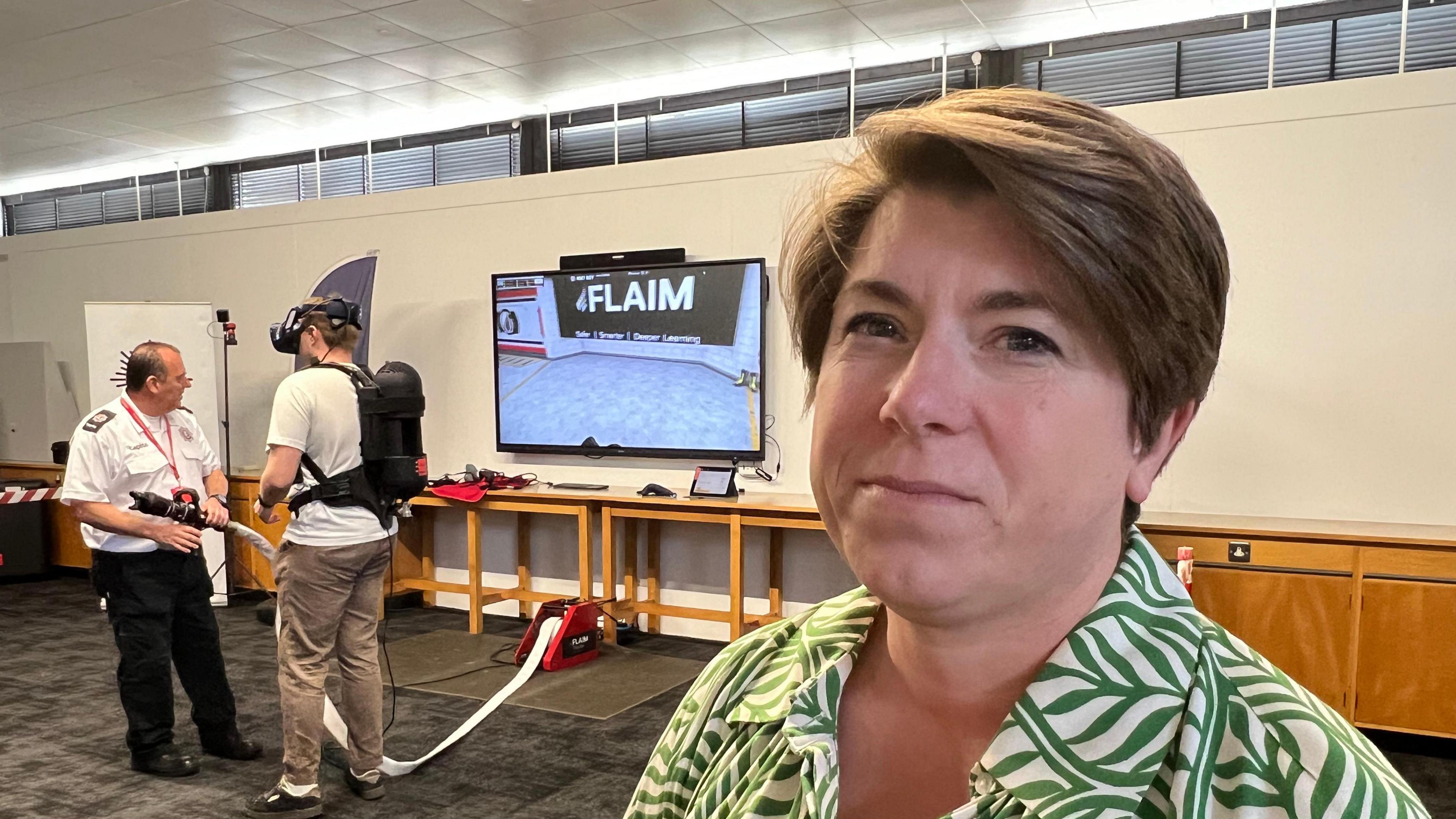 A woman wearing a green and cream patterned shirt standing in front of virtual reality TV screen and equipment. 