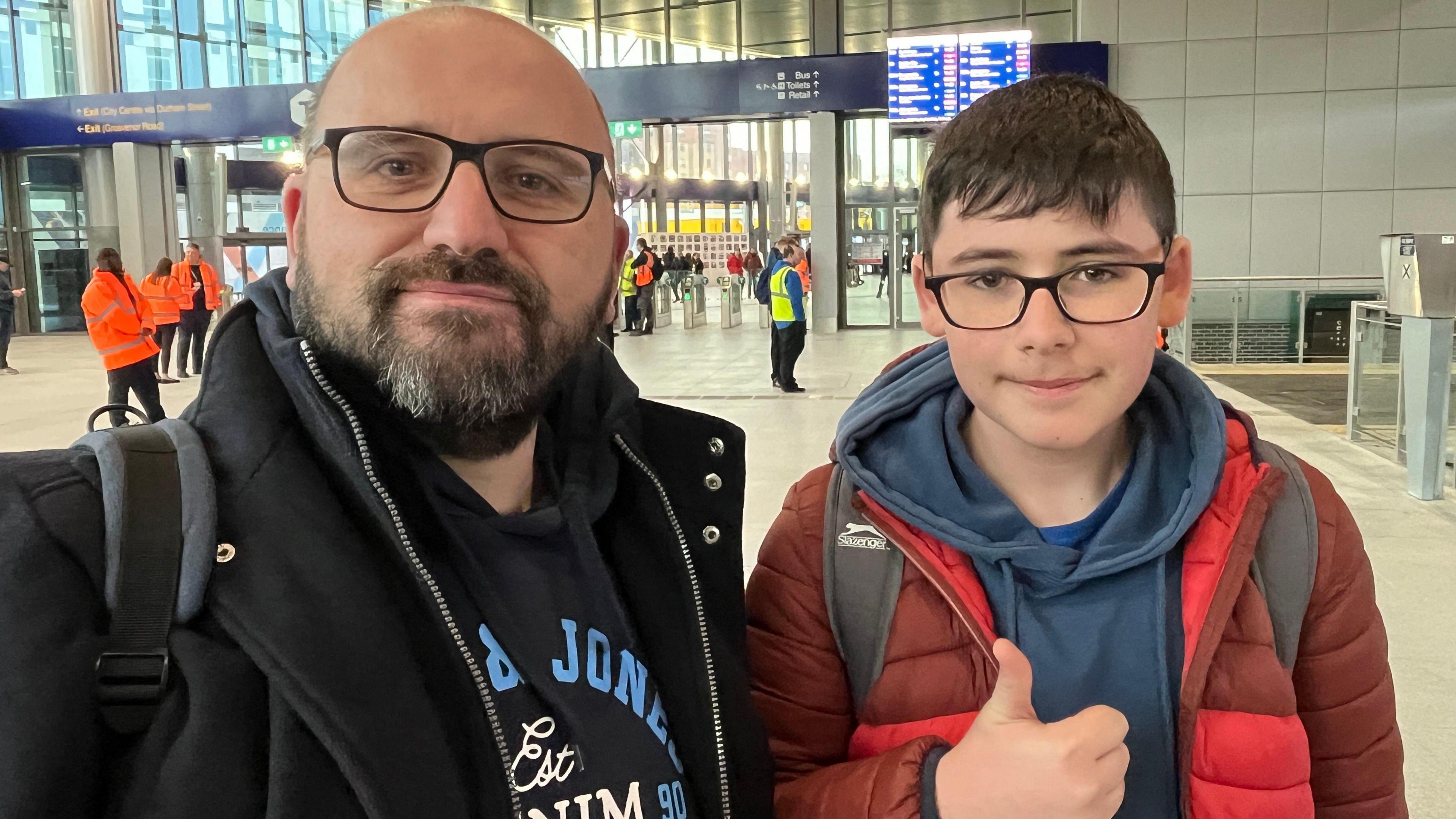 A man and a boy look into the camera. The man, who's on the left, wears black glasses and a black coat. His son, who's on the right, also wears black glasses and is giving a thumbs up sign to the camera. He's wearing a red coat with a grey hoodie underneath.