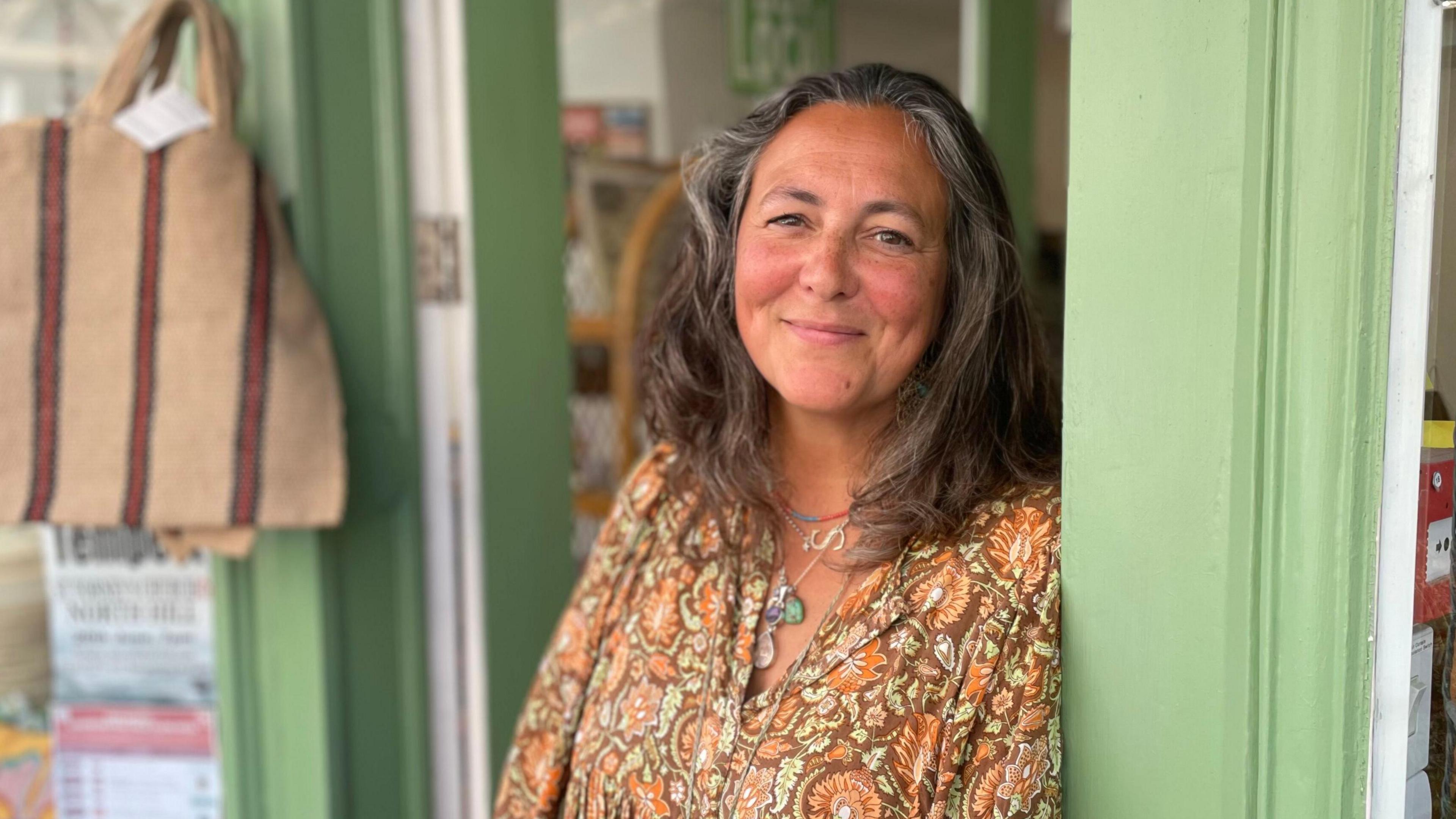 A woman leans on a shop doorway, smiling at the camera. She has shoulder-length hair and is wearing a brown and yellow patterned top and several necklaces
