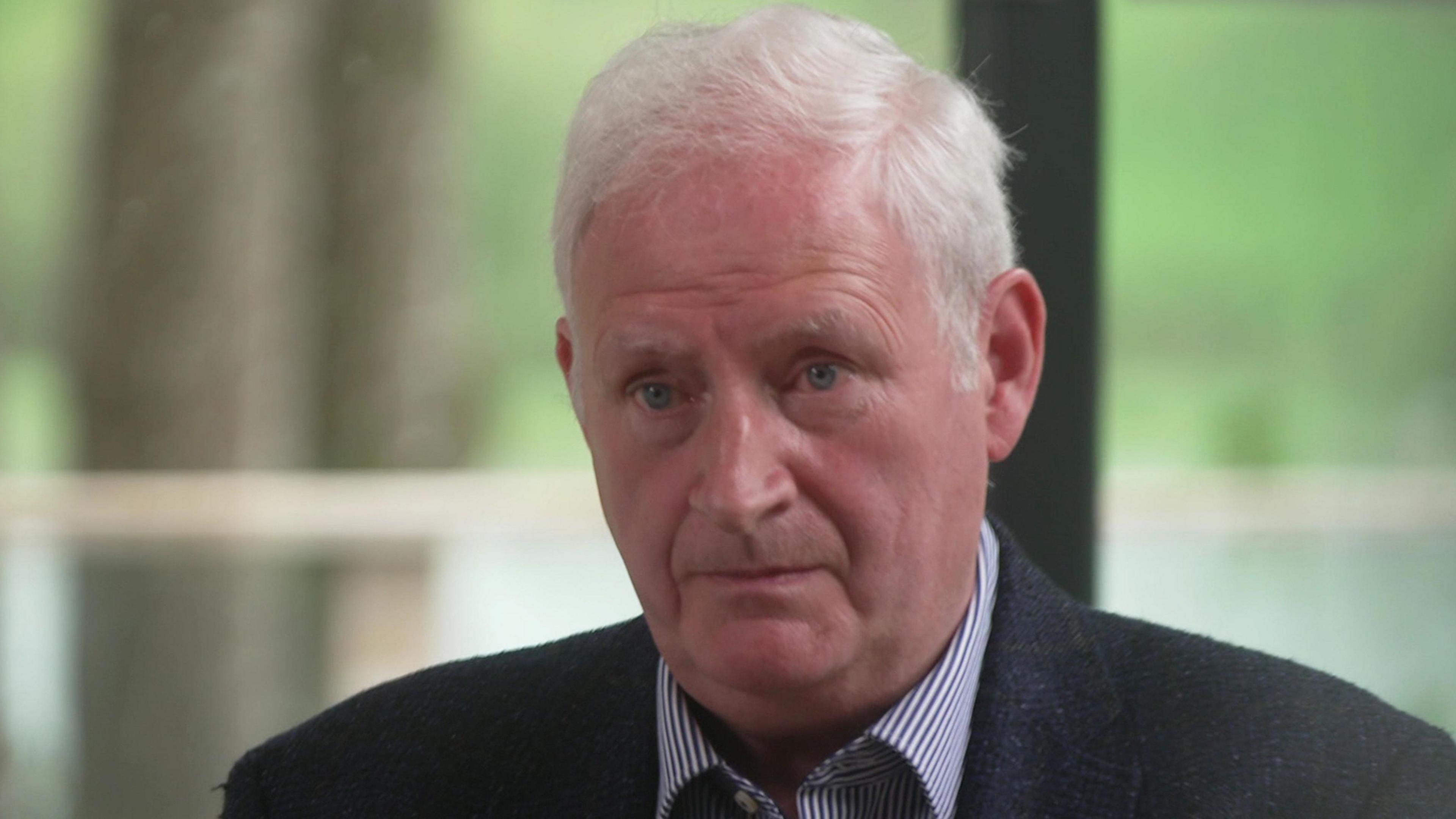 A man with short grey hair, which is brushed to the side, is sitting in front of a large window or door, with a blurred background of grass and a tree trunk. He is wearing a blue and white pinstriped shirt and a dark jacket. 