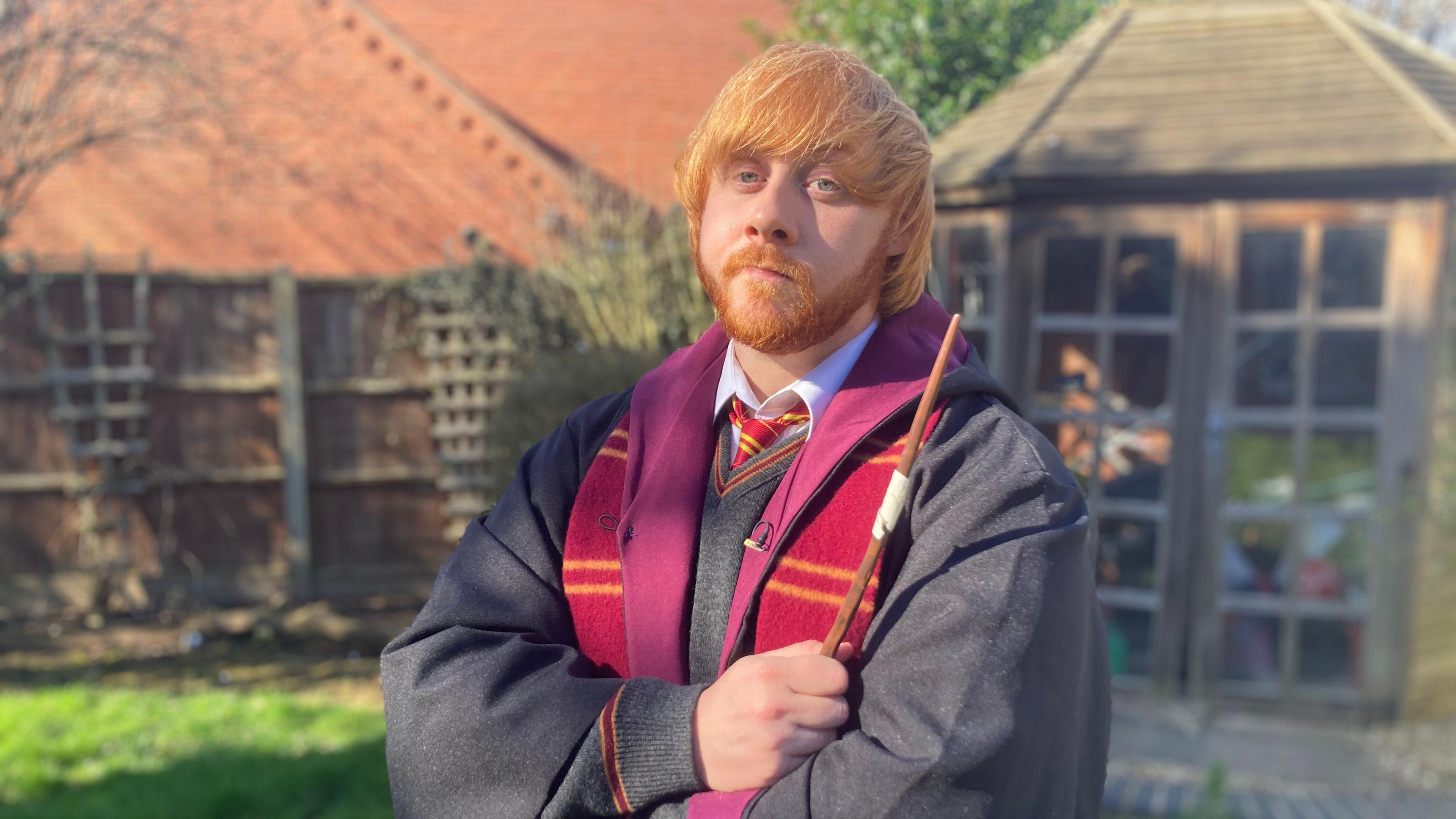 A man with ginger hair which is swept across his forehead and a ginger moustache and beard pulling a serious pose for the camera. He is wearing a Hogwarts school uniform with his arms crossed and a wand is in his hand.