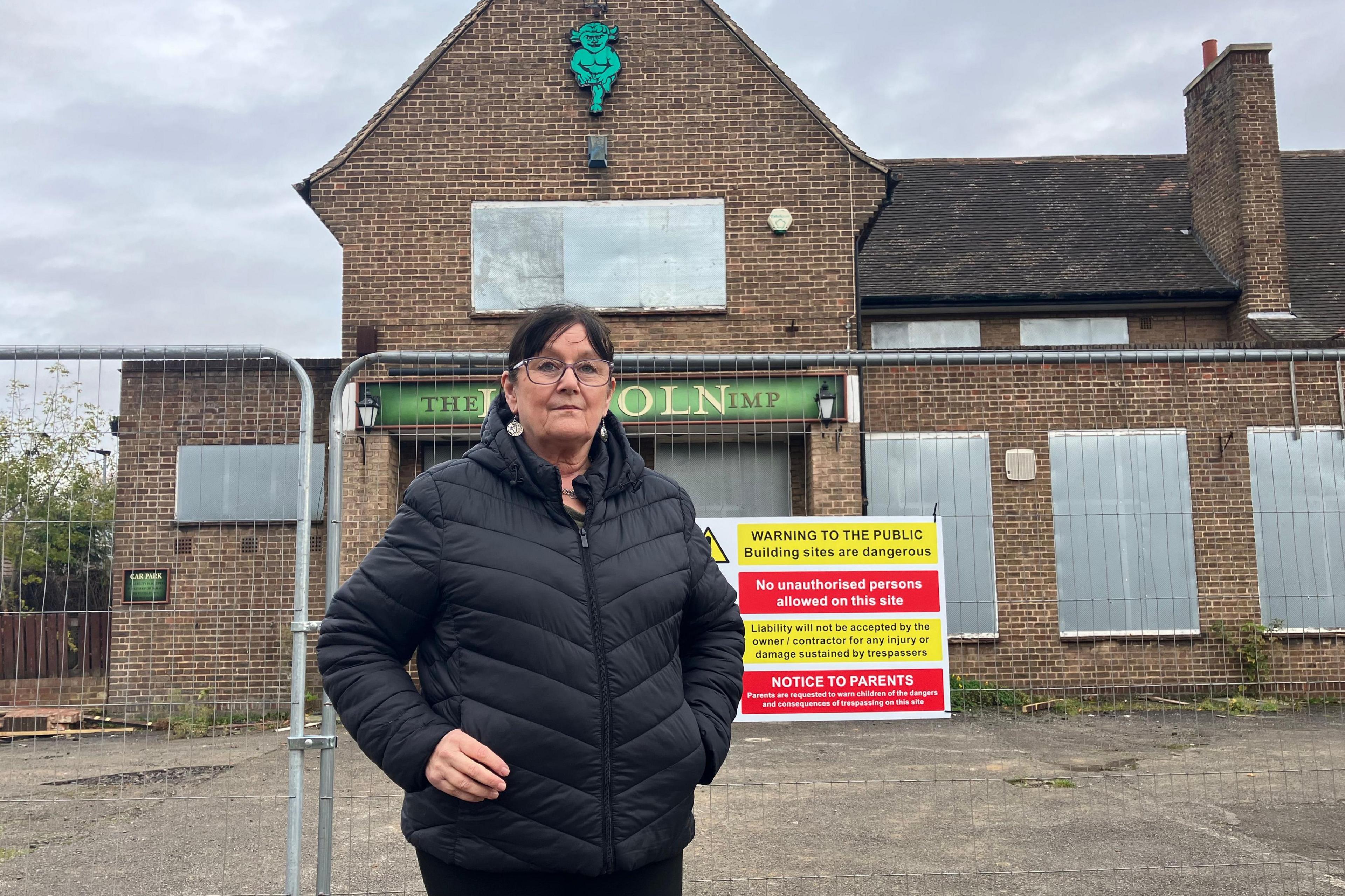 A woman with brown hair and glasses and a dark jacket stood in front of front of a derelict pub.