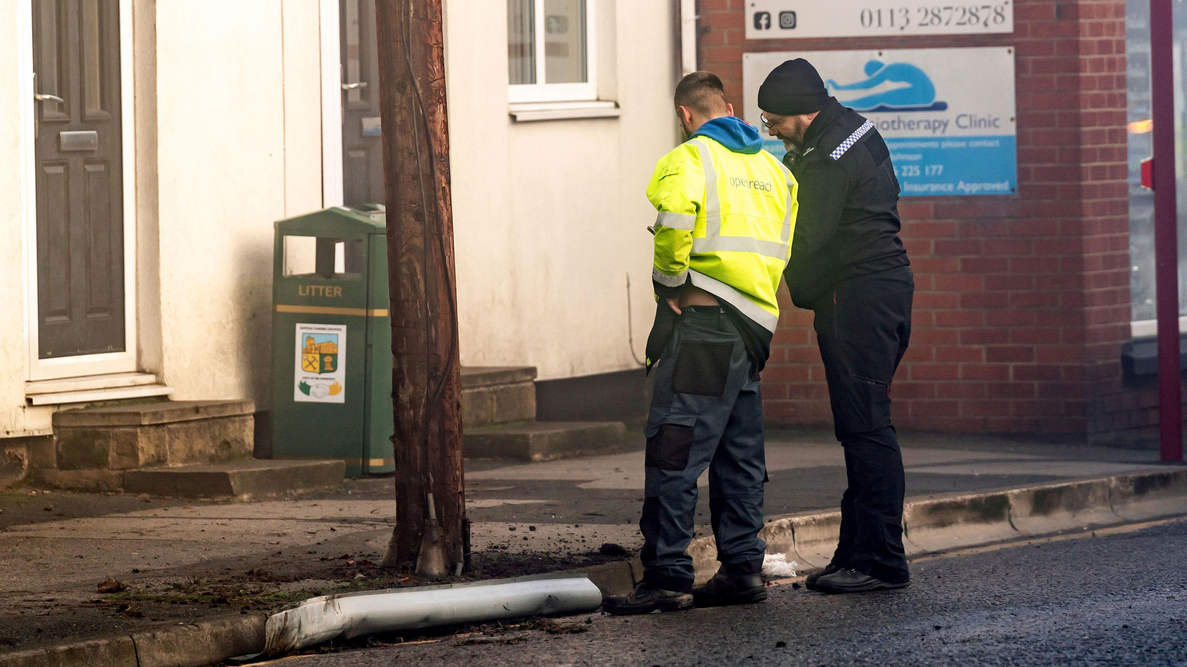 A police officer and BT Openreach worker stand next to a telegraph pole in High Street, Kippax. The remnants of a fire can be seen on the ground.