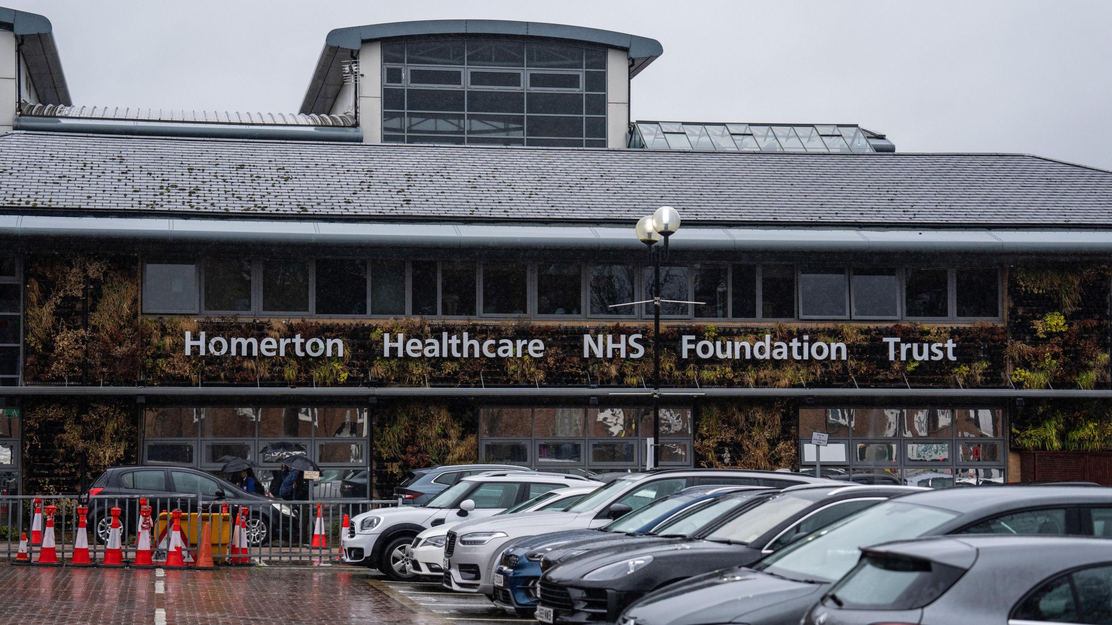 The facade of Homerton Hospital covered in foliage and a row of windows. The wording Homerton Healthcare NHS Foundation Trust is in large white writing along the front of the building. 