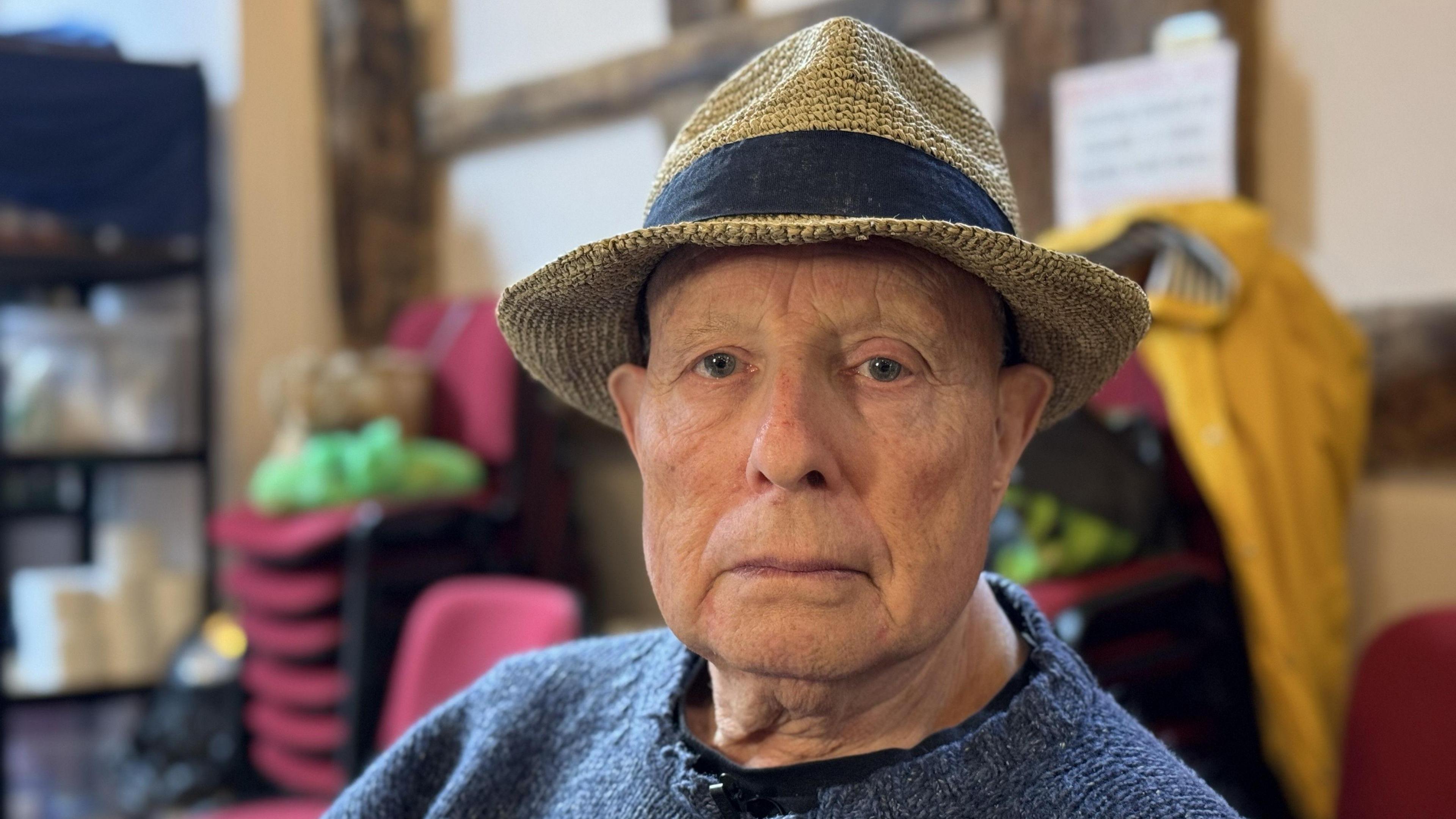 A man with a beige boater hat with a navy stripe above its brim. He is wearing a navy jumper and there are red chairs stacked behind him