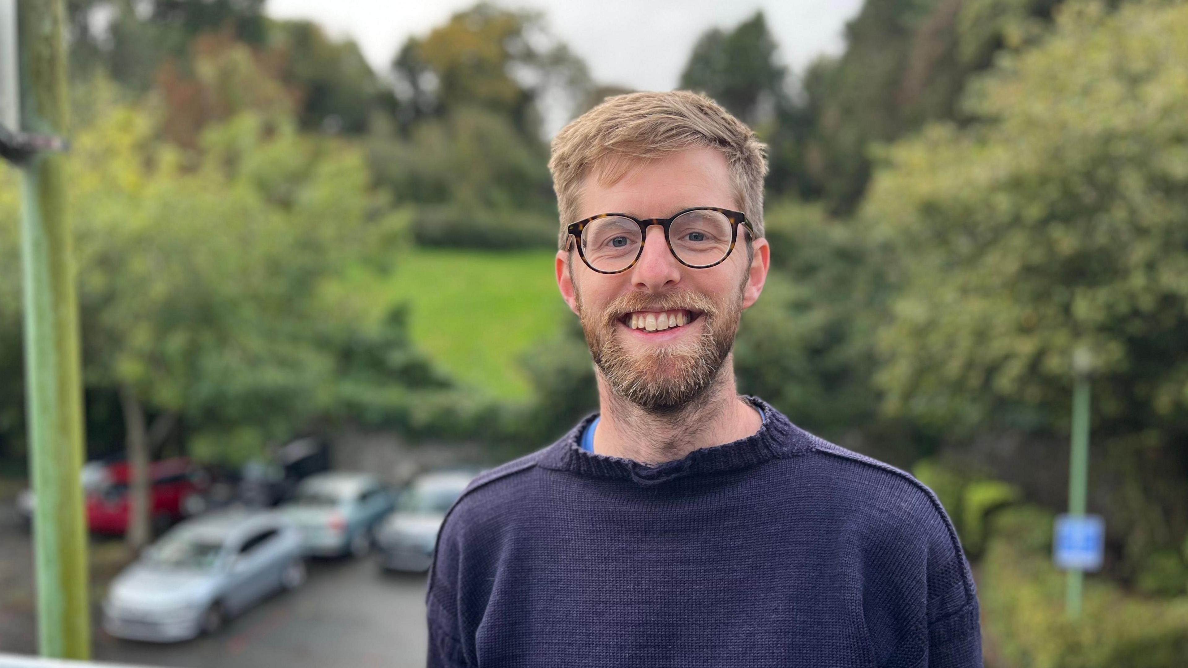 A man with blonde hair and glasses, with a blonde beard smiling towards the camera. He's also wearing a blue Guernsey jumper with a blue t-shirt underneath. 