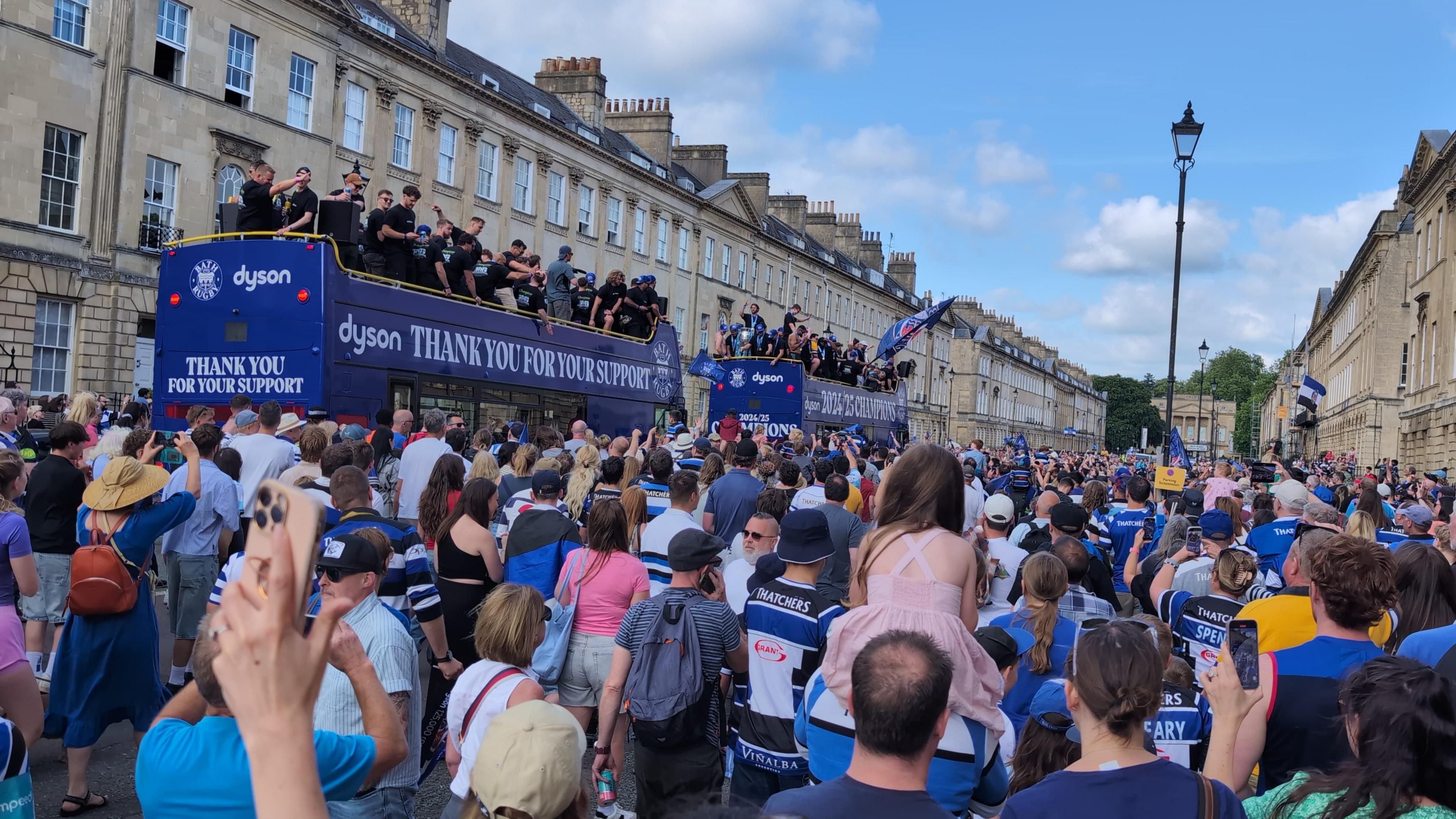 Fans in blue black and white waving flags with two blue open top buses passing in the background with players on top waving