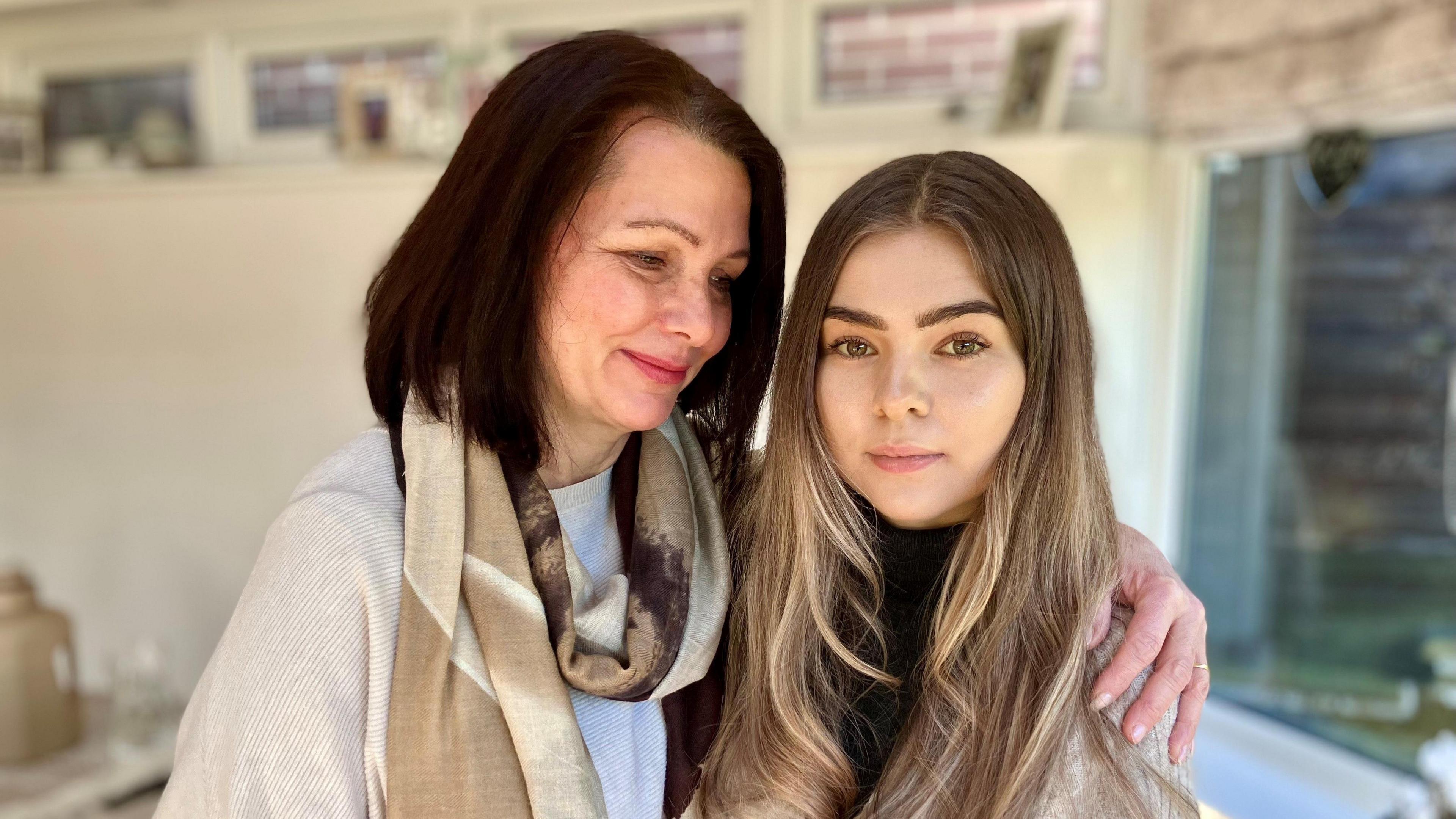 A middle aged, white woman with brown hair holds a younger woman in her arms and looks at her fondly.  They are standing in a home by a window. The younger woman has lighter brown hair and brown eyes.