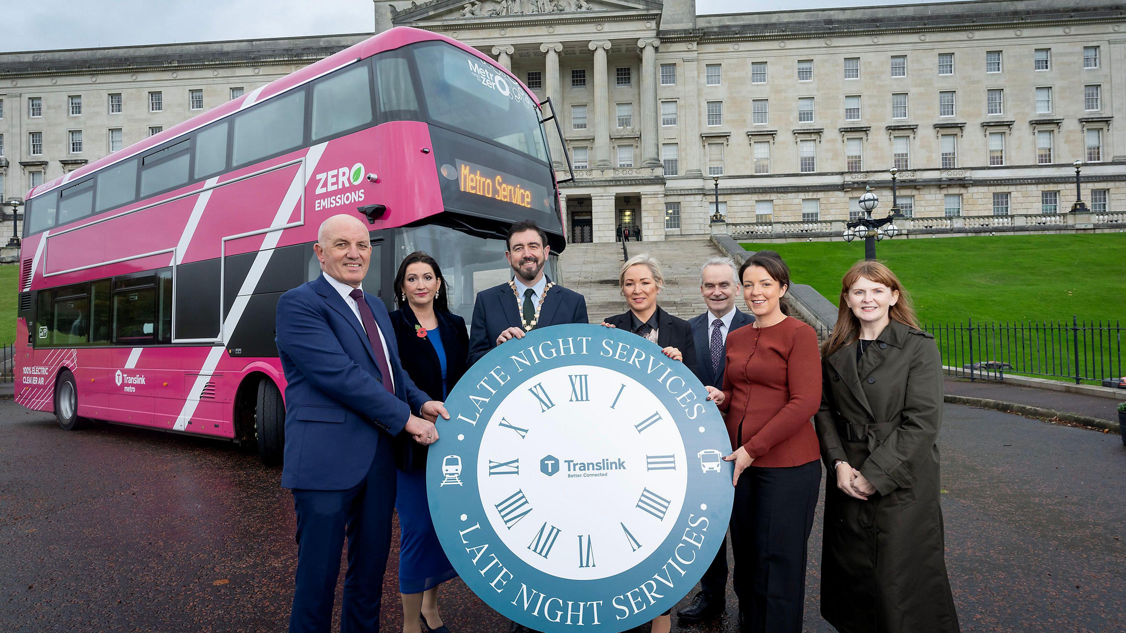 Seven people pictured with a blue and white clock in front of a pink Translink bus. They are in front of Stormont.