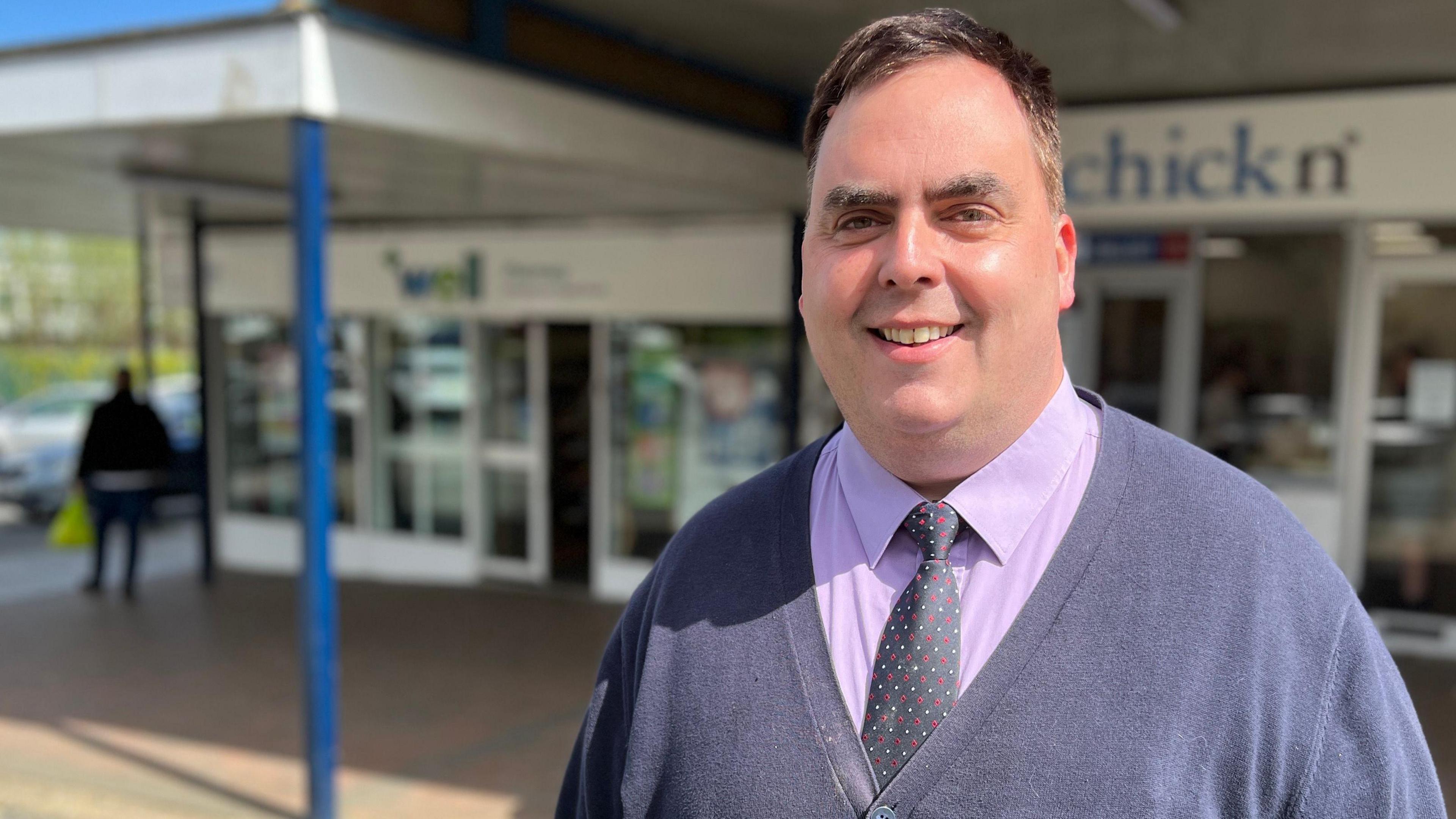 Kerry Smith wearing a cardigan, purple shirt and spotted tie. His hair is dark brown. He is smiling. He is standing outside in front of a parade of shops.