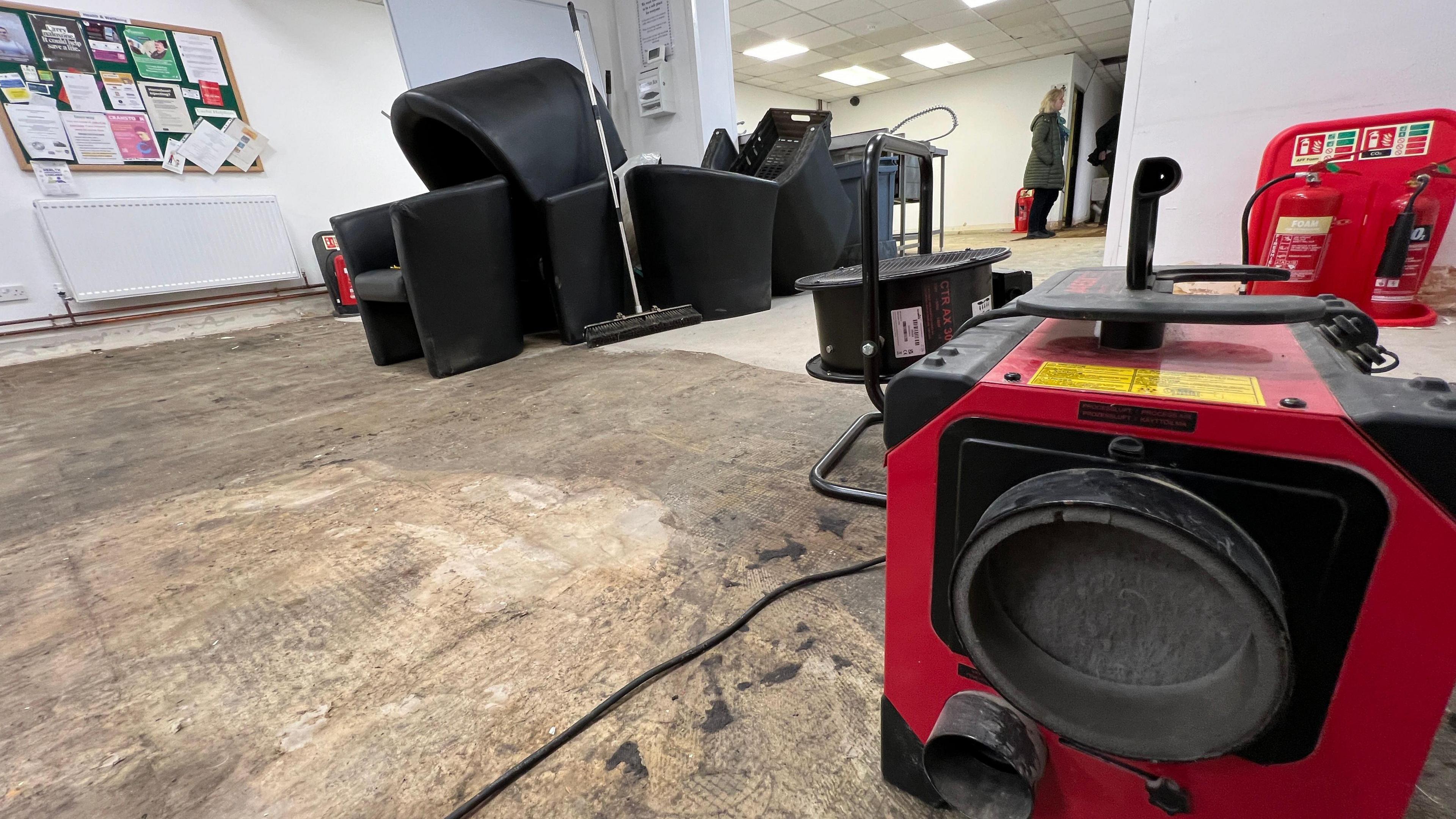 A large red industrial fan in the middle of the hall at The Citadel. Some of the floor boards have been removed revealing the flooring underneath.