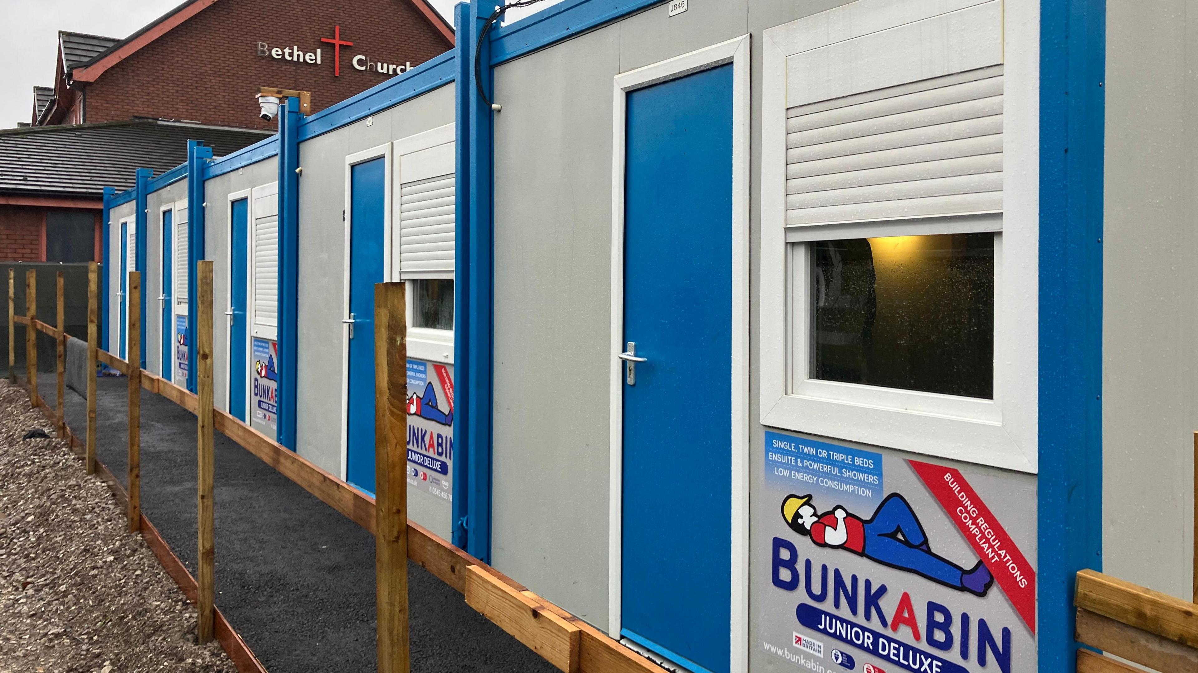 Five of the sleeper pods stretching into the distance. Each has a blue door and a window with white shutter. 