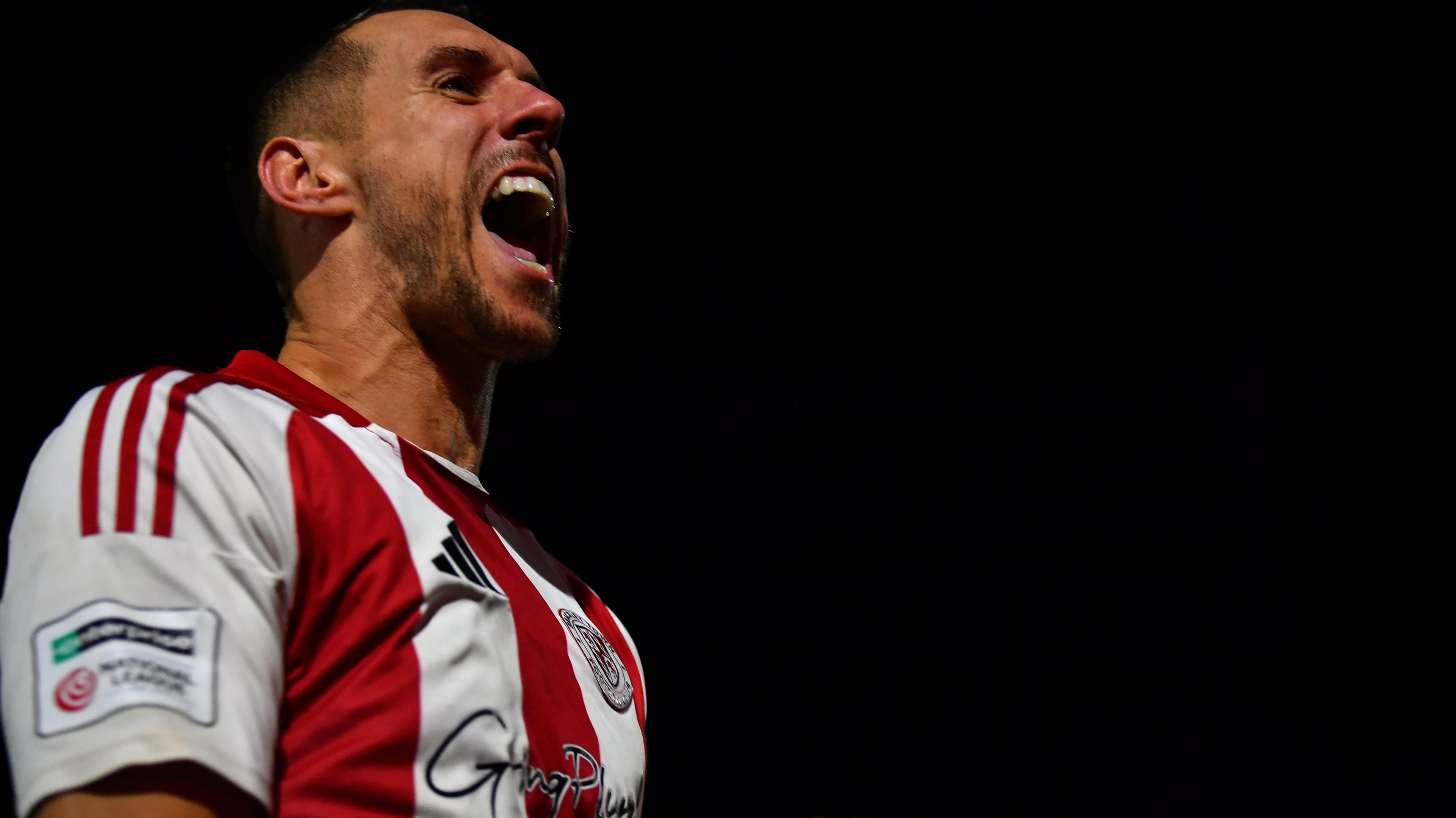 A photo of footballer Matt Lowe celebrating a goal while playing FA Cup football for Brackley Town at Notts County in the FA Cup. Lowe has his mouth open and is looking to his left against a night sky while wearing a red and white striped shirt with a black Adidas logo and the club shirt visible on the chest