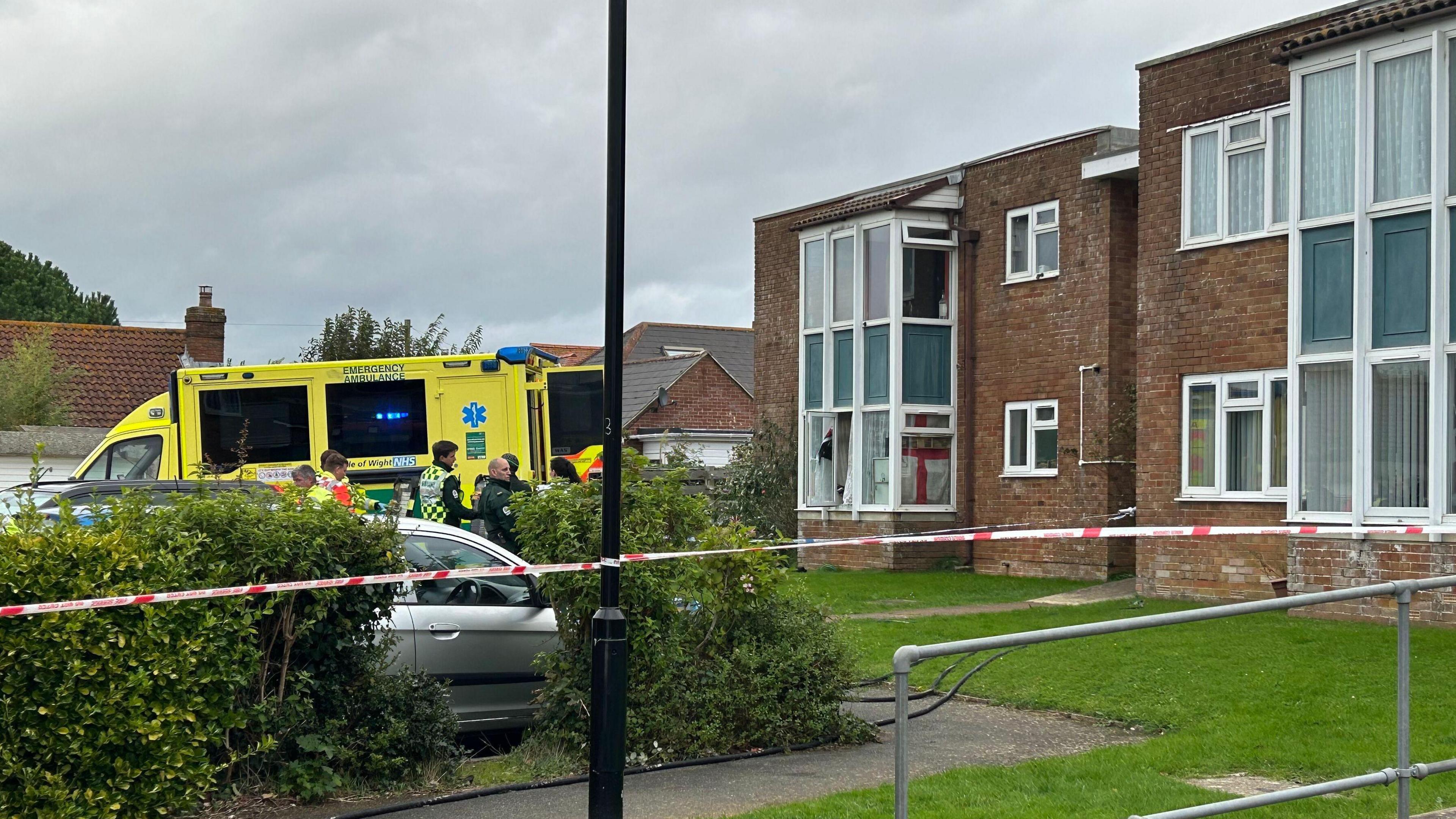 A two-storey block of flats with a grass verge in front of it, cordoned off with red and white tape. An ambulance is stopped near the property, with paramedics and other emergency workers milling around.